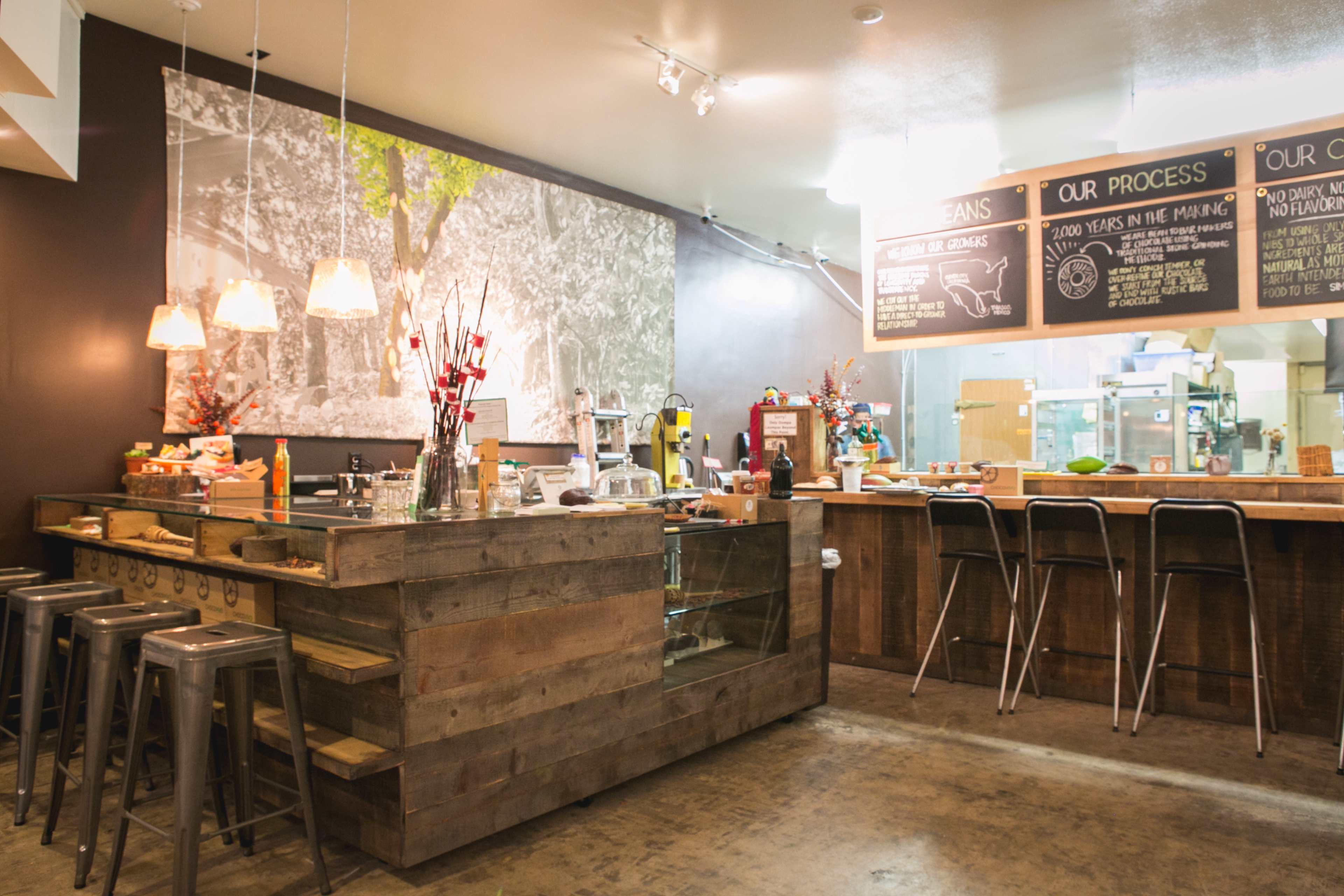 A rustic café interior with a wooden counter, metal barstools, and a large blackboard detailing the menu and process against a backdrop of a wall mural.