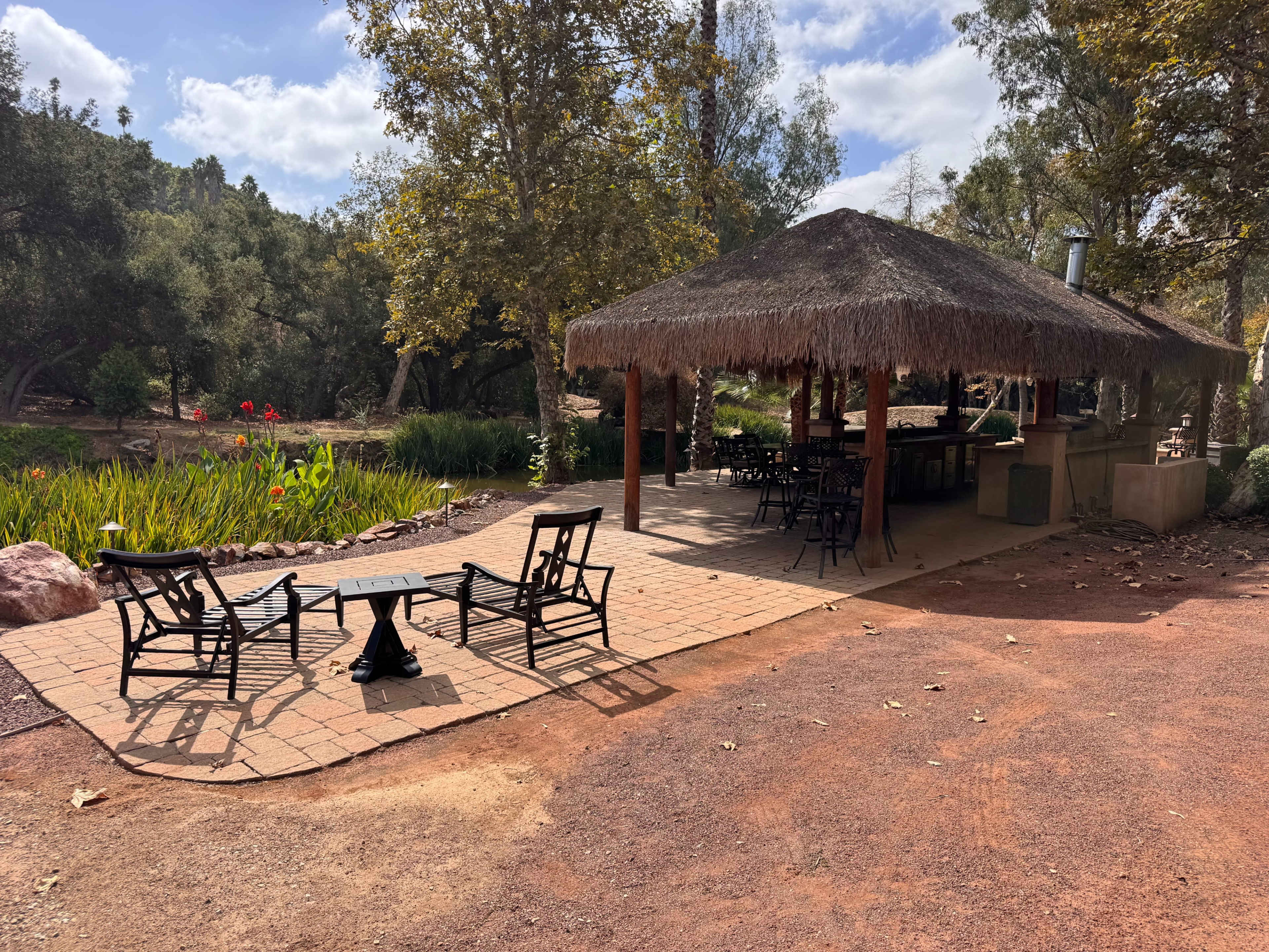 A thatched roof outdoor seating area with wooden chairs and a small table is situated near a landscaped garden.