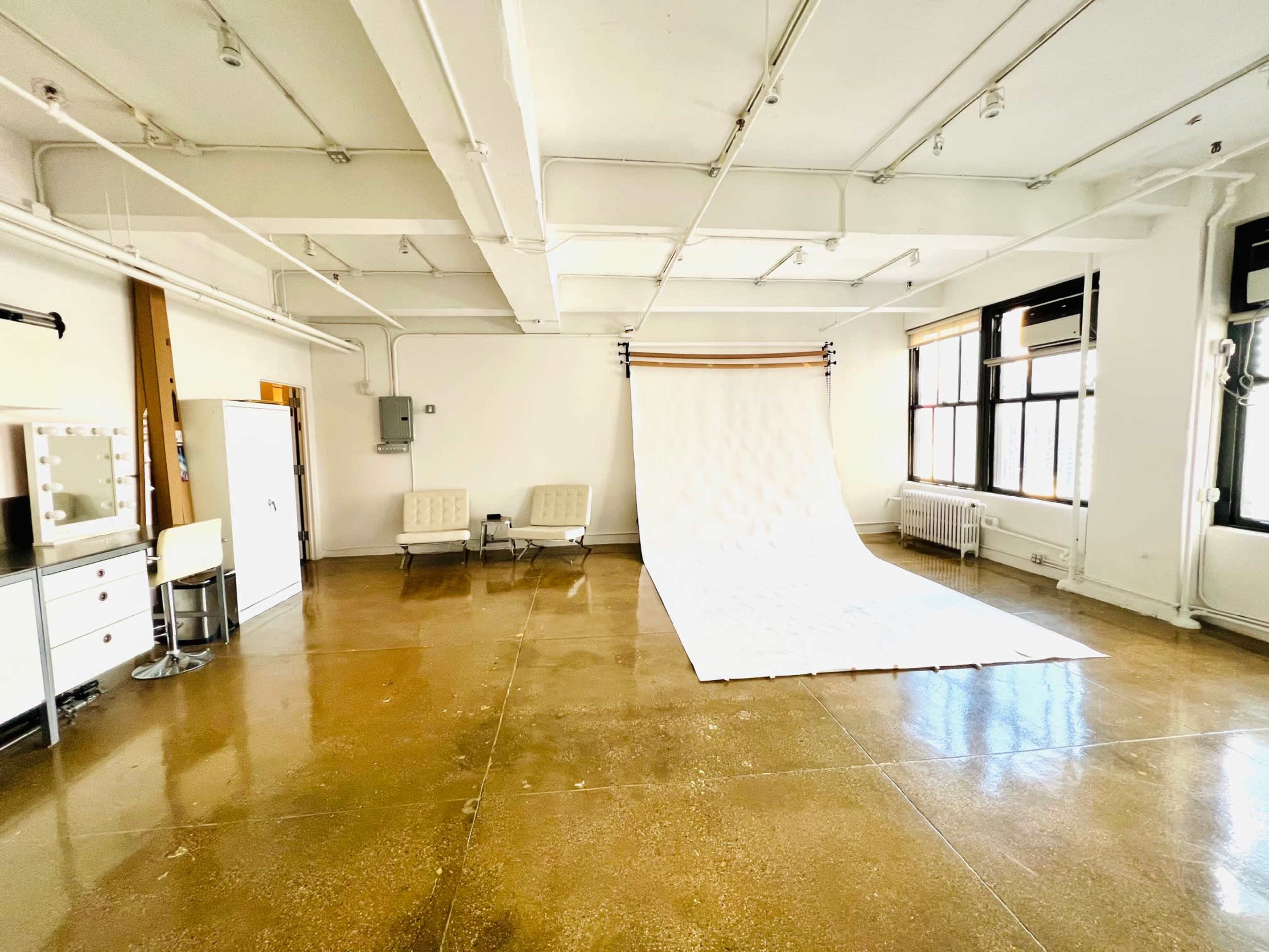 A spacious studio with a glossy concrete floor, a white backdrop, and a seating area featuring two chairs and a vanity with mirrors.