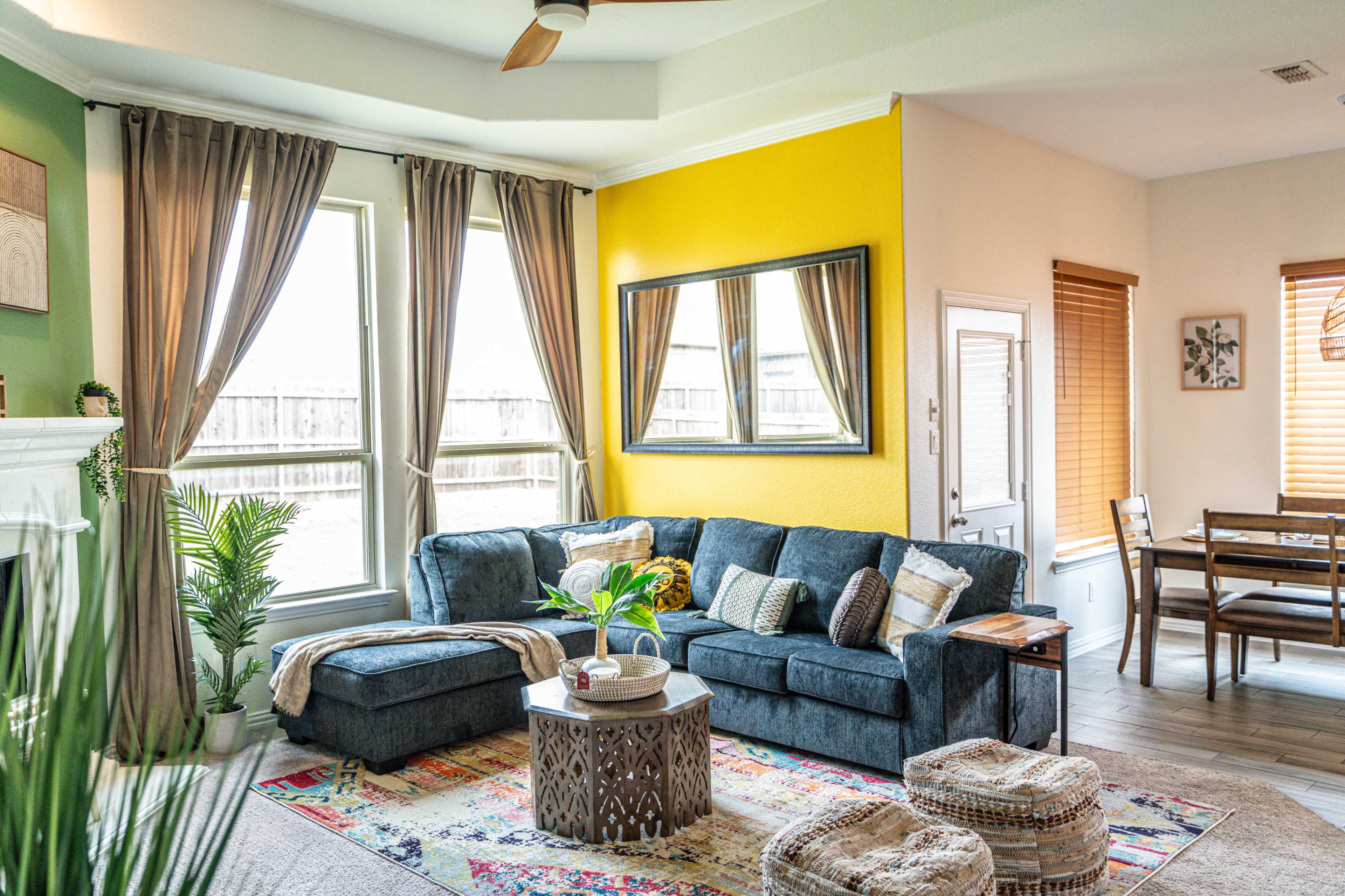 A modern living room features a sectional sofa, decorative pillows, a round coffee table, a large mirror, and a combination of natural light and vibrant wall colors.