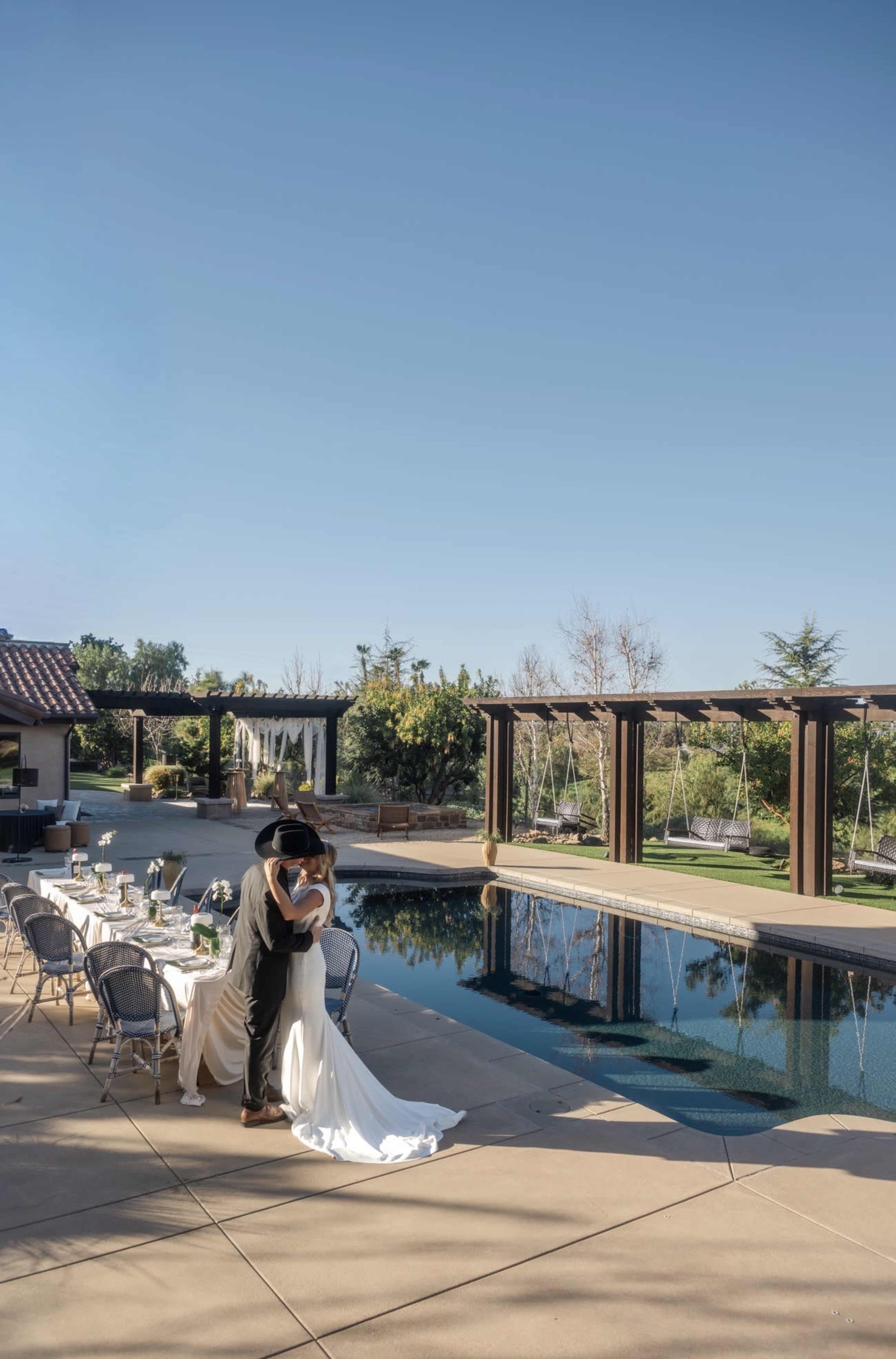 A couple embraces near a poolside dining area adorned with a table set for an outdoor wedding reception.