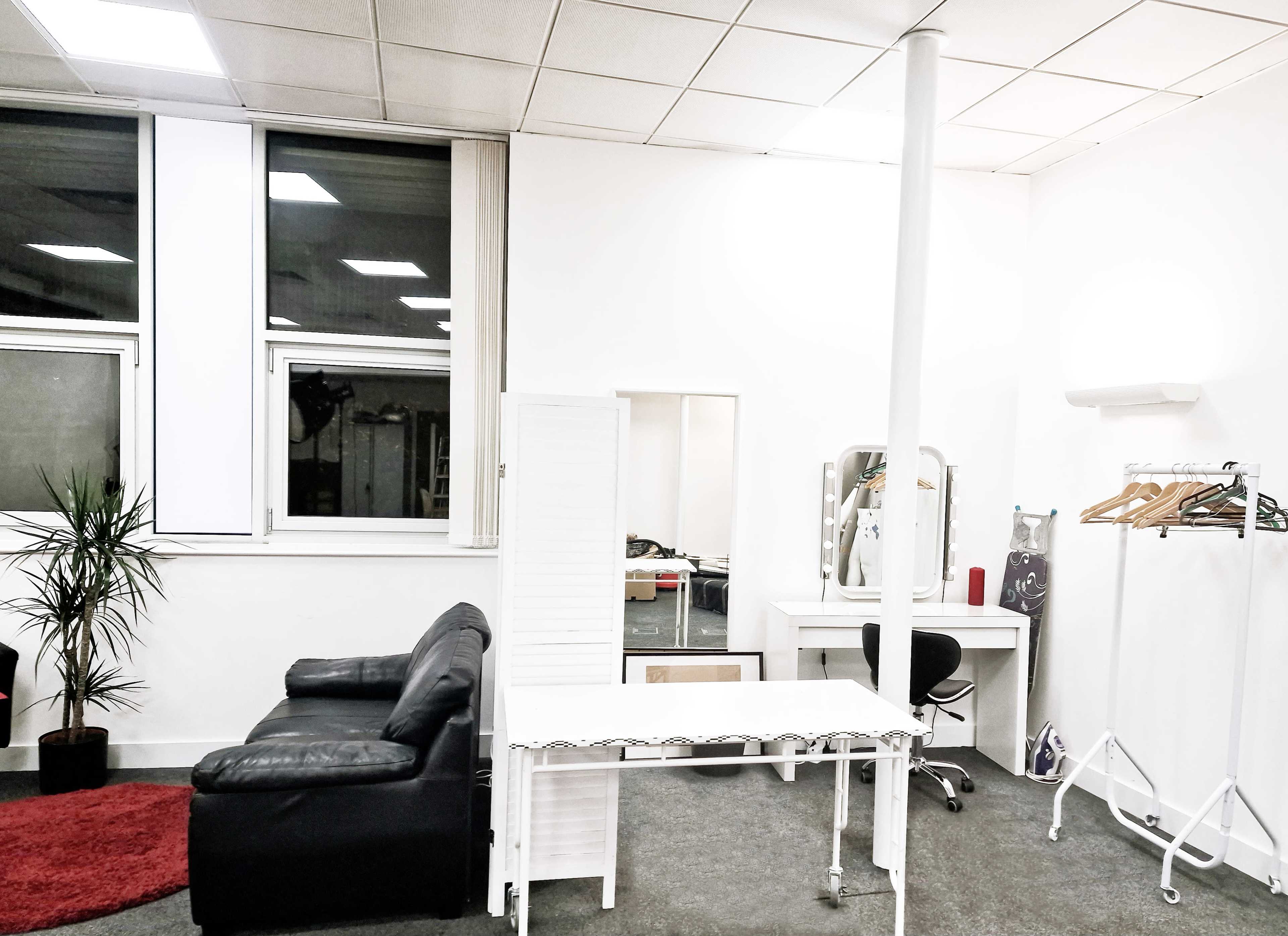 An office space featuring a black couch, a white dressing table with a mirror, a clothing rack, and a small plant beside a window.