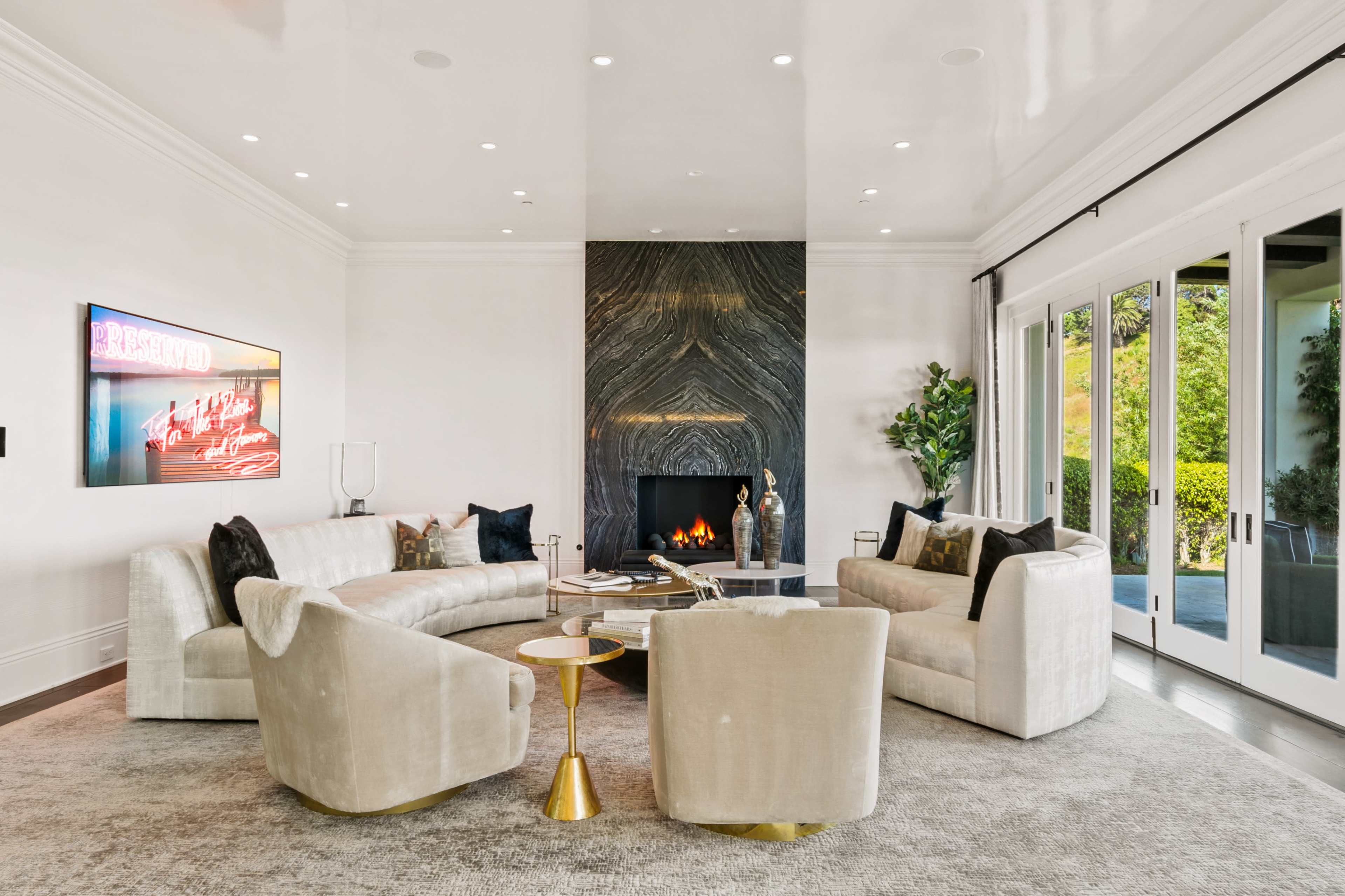 The image shows a contemporary living room featuring a curved sofa, two armchairs, a fireplace with a dark stone surround, and large glass doors leading to the outdoors.