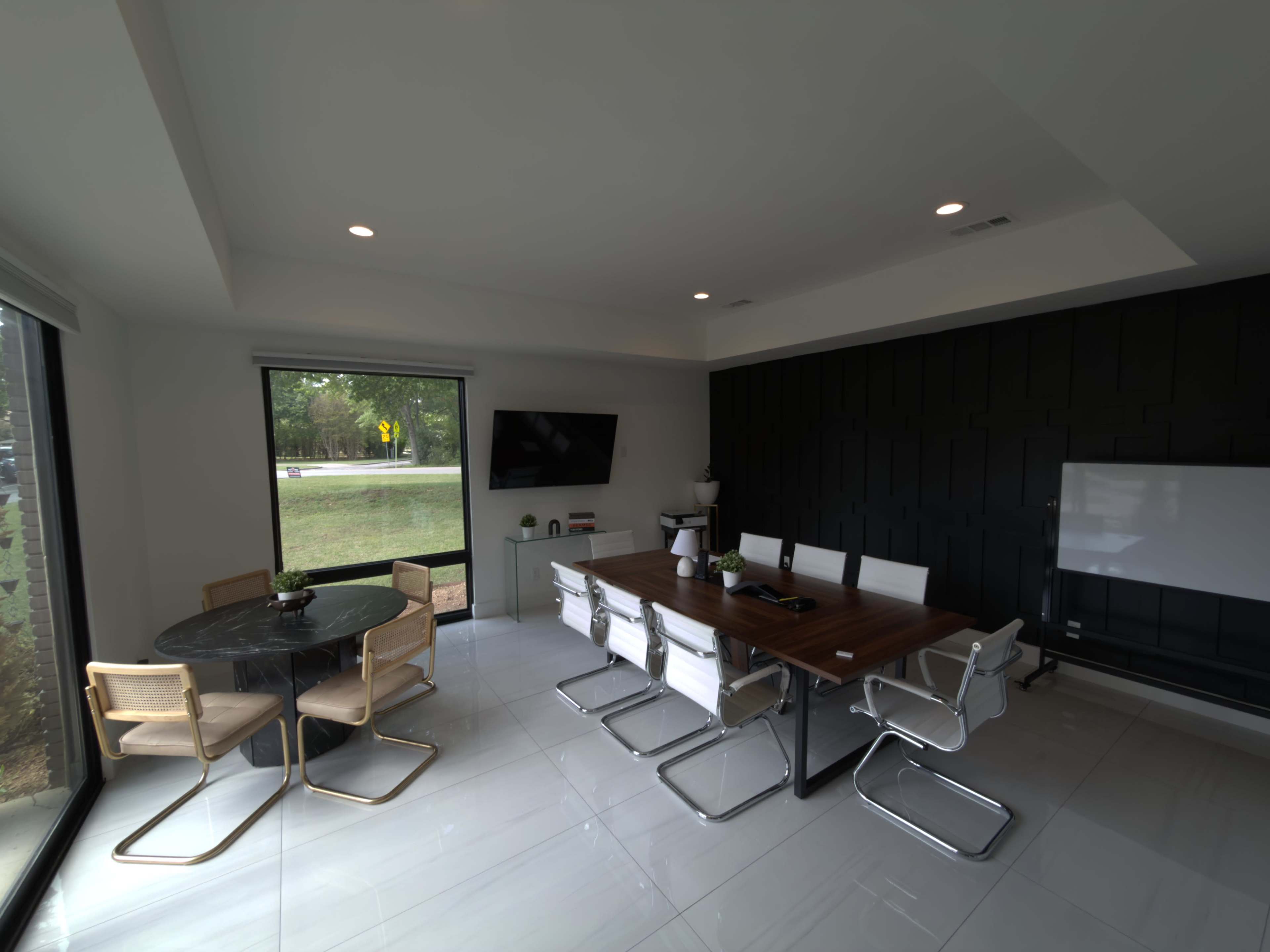 The image shows a modern conference room with a large rectangular table, white chairs, a circular table in the corner, a television on the wall, and large windows providing outdoor views.