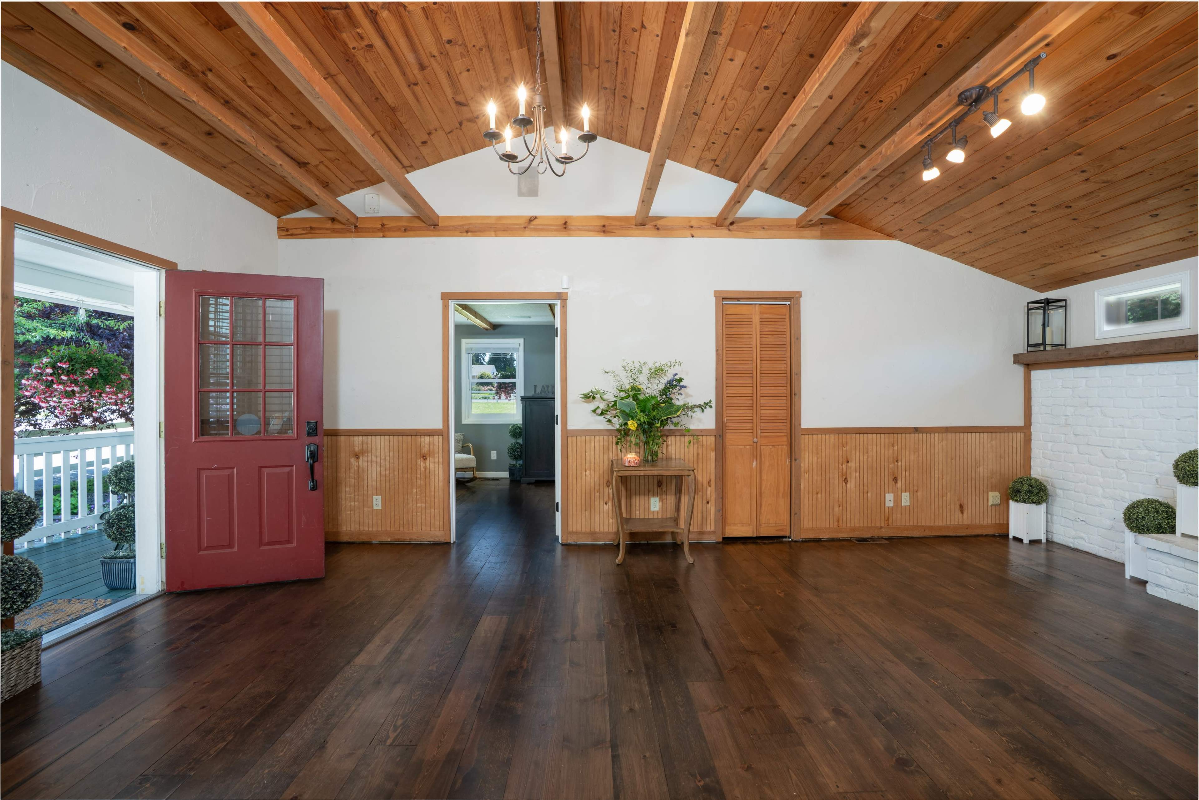 The image shows a room with wooden ceiling beams, a red front door, two doorways leading to other spaces, and decorative plants in the corners.