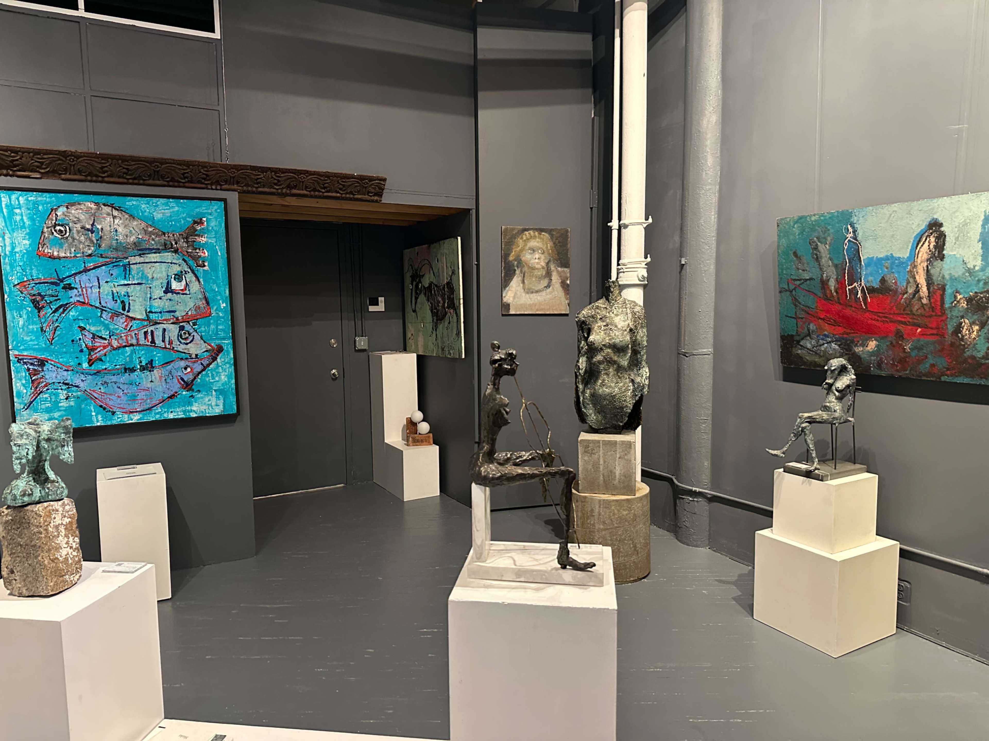 The image shows an art gallery featuring various sculptures and paintings displayed on gray walls, with installations on white pedestals and one large colorful painting of fish.
