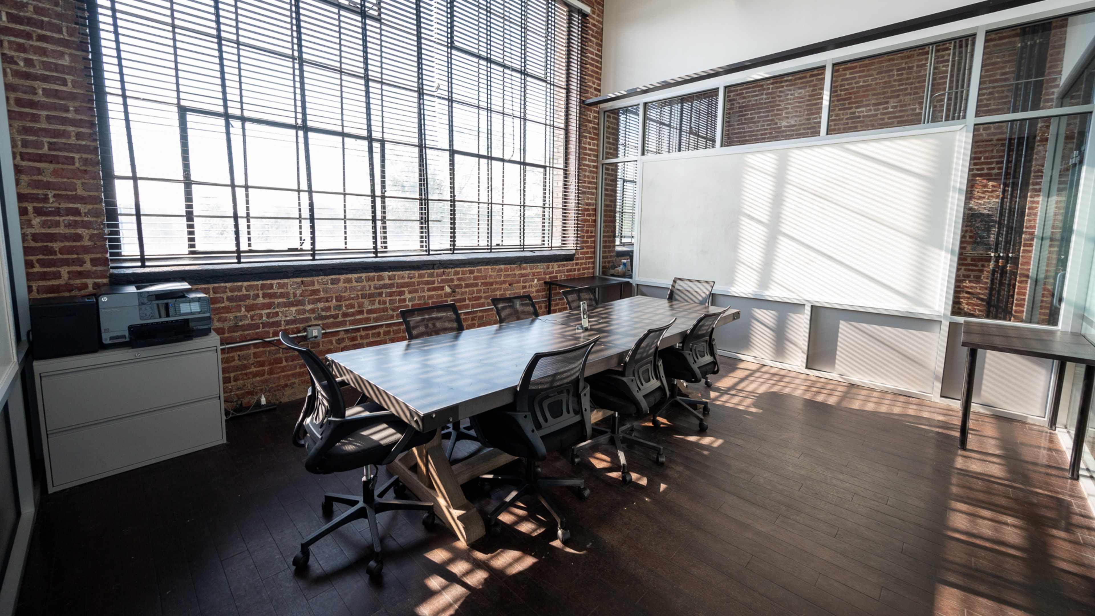 A conference room features a long table surrounded by chairs, with large windows and exposed brick walls.