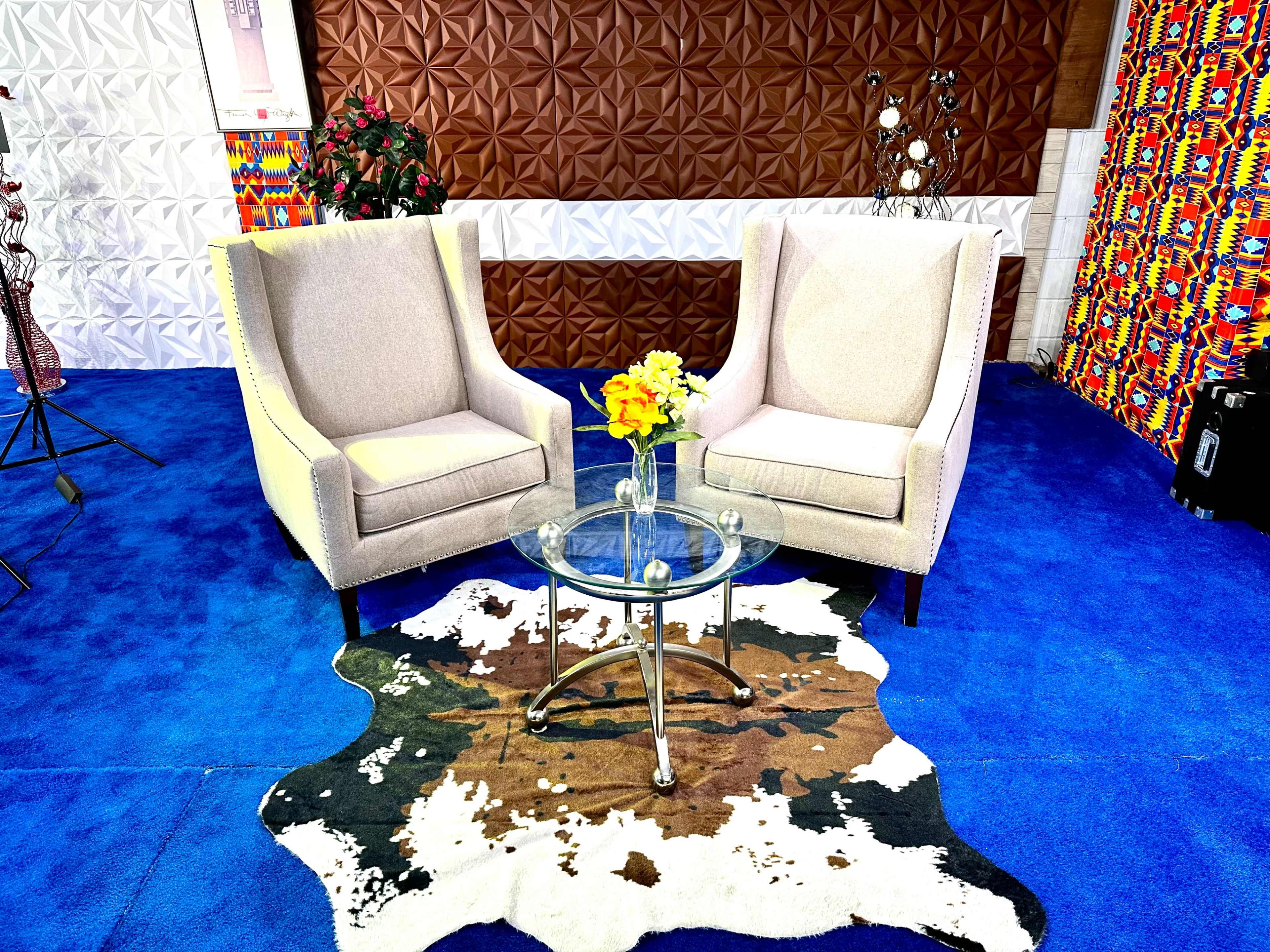 Two beige armchairs face each other across a glass coffee table, situated on a cowhide rug in a room with colored patterned walls.