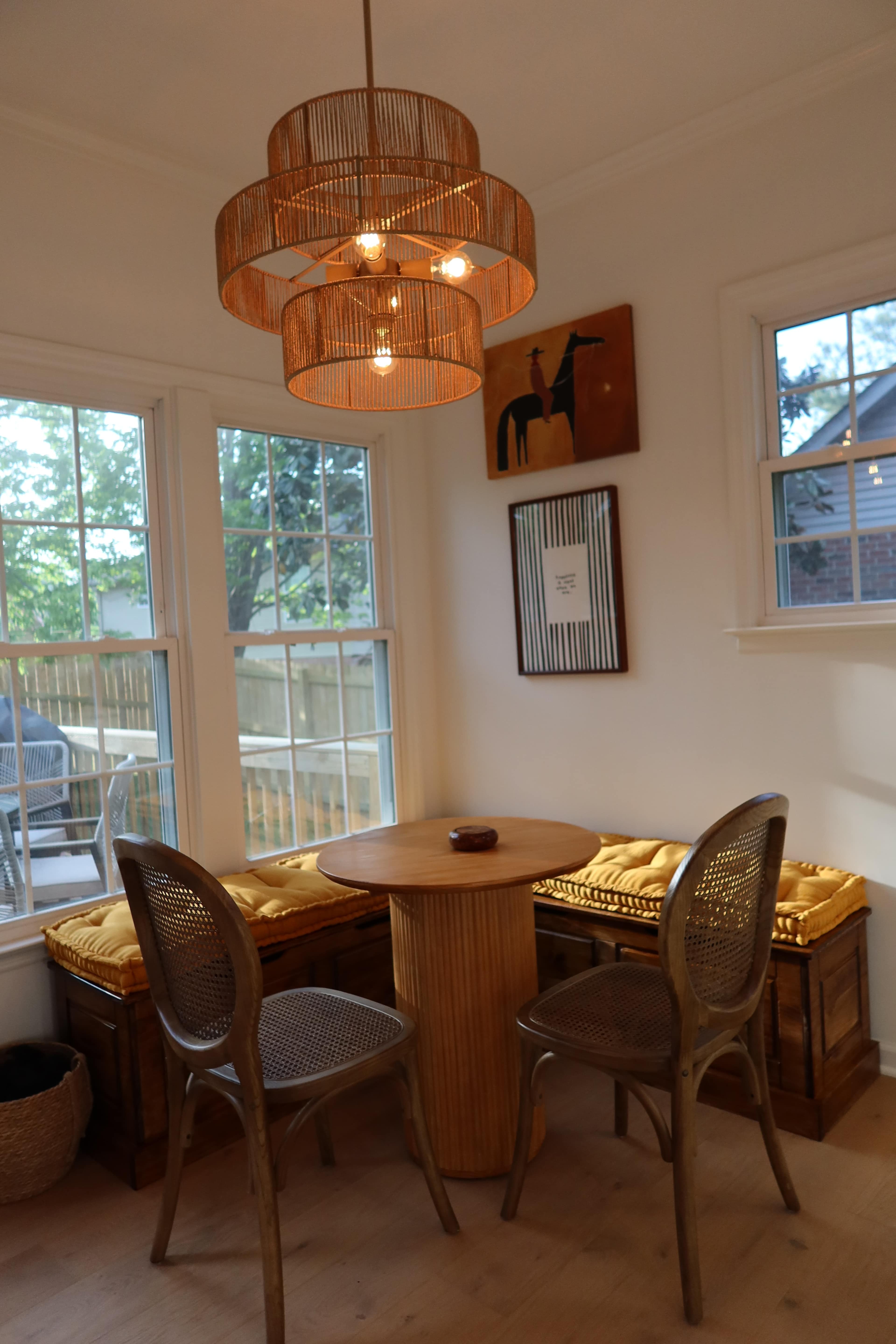 The image shows a dining area featuring a round wooden table, two chairs, and a built-in bench with cushions, illuminated by a large pendant light.
