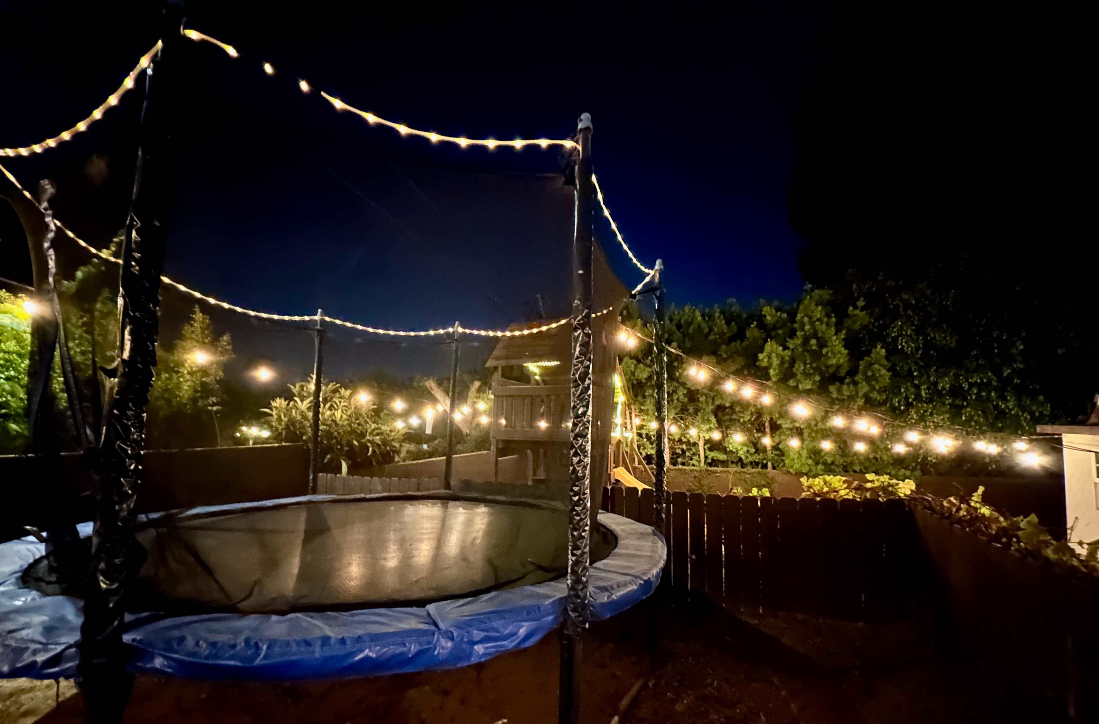 A trampoline with blue padding is illuminated by string lights in a backyard at night.