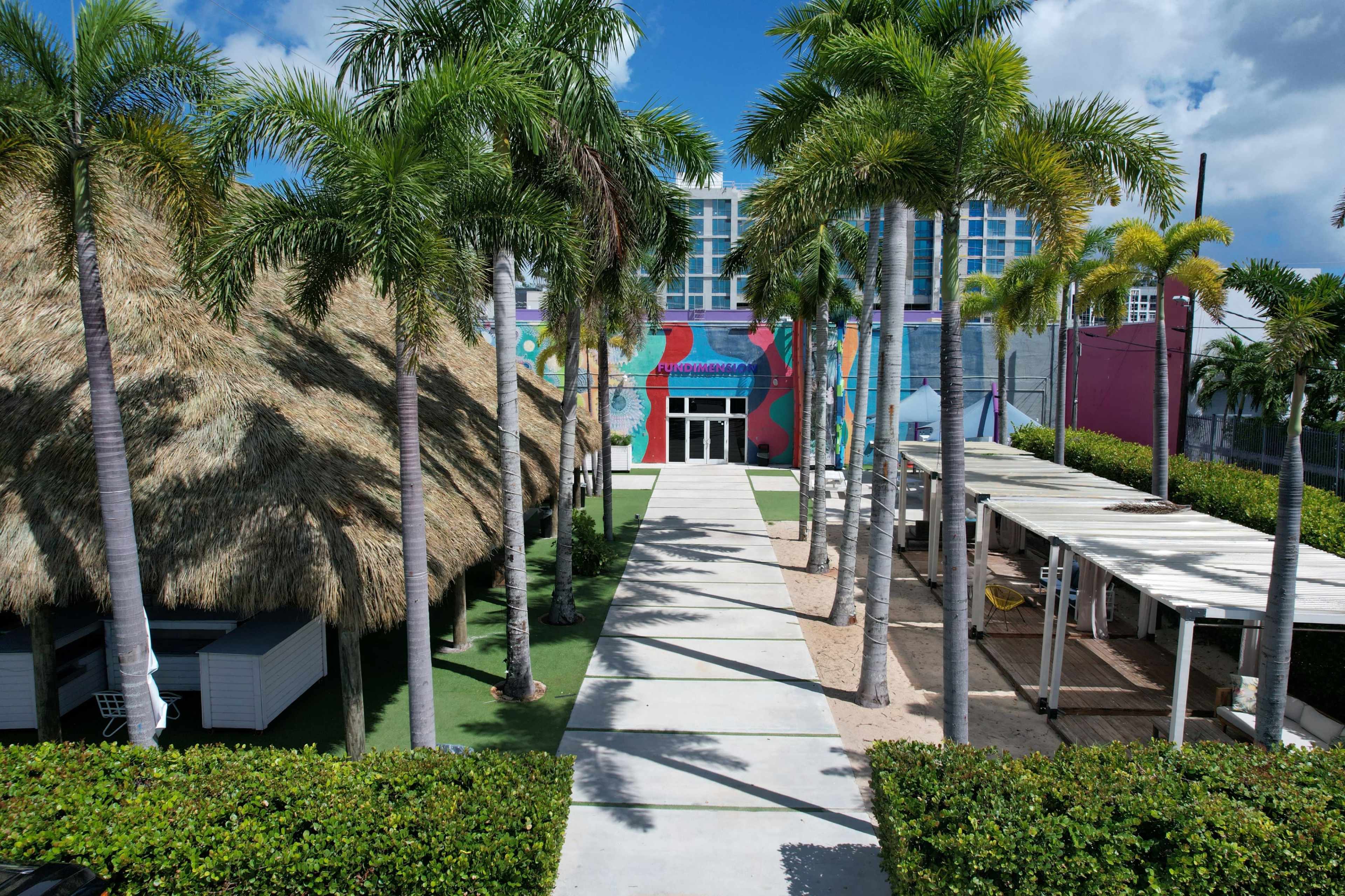 The image shows a pathway lined with palm trees leading to a colorful building with a large mural, flanked by thatched and open structures.