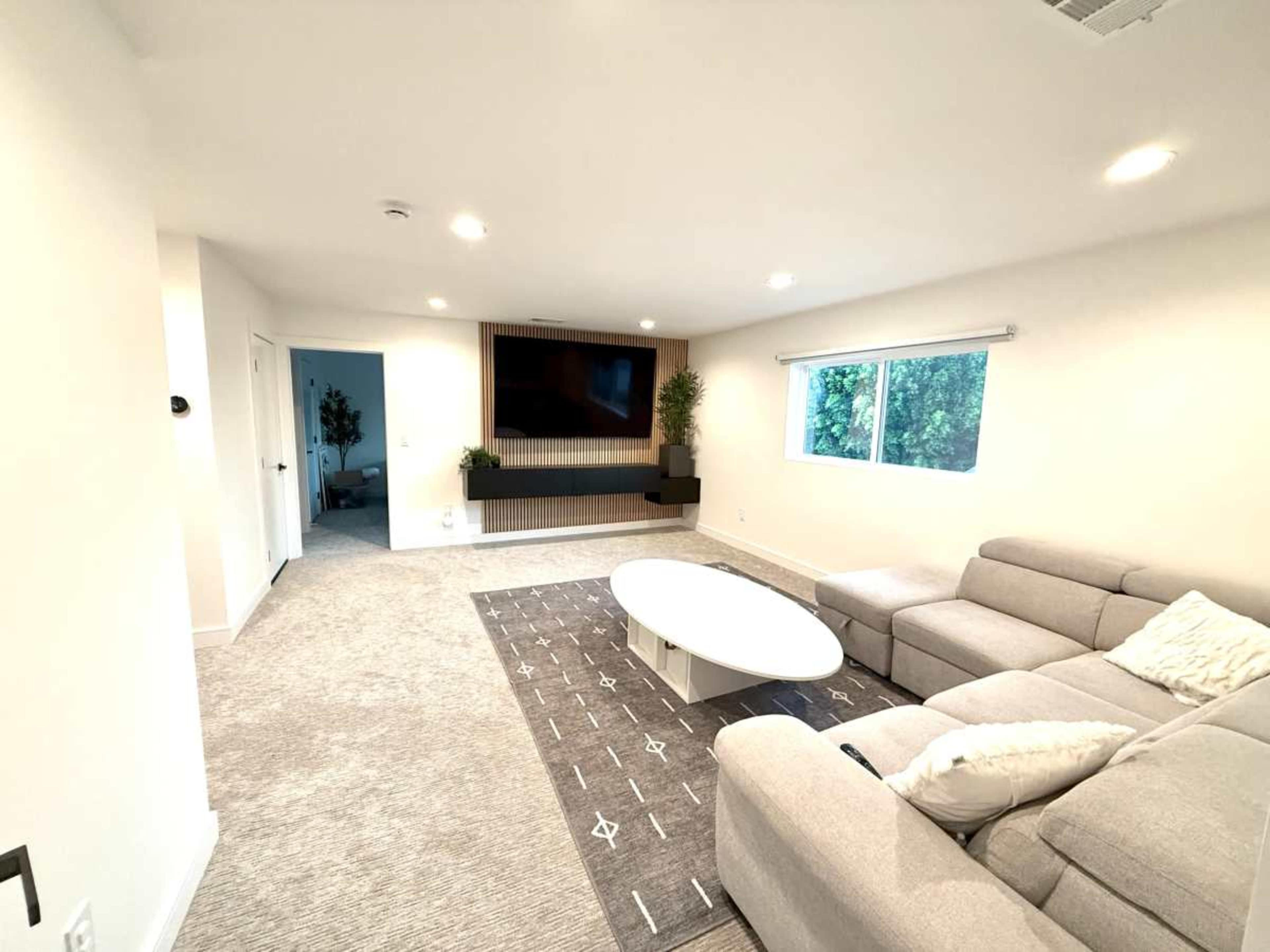 The image shows a modern living room with a light gray sectional sofa, a round white coffee table, and a large flat-screen TV mounted on the wall.