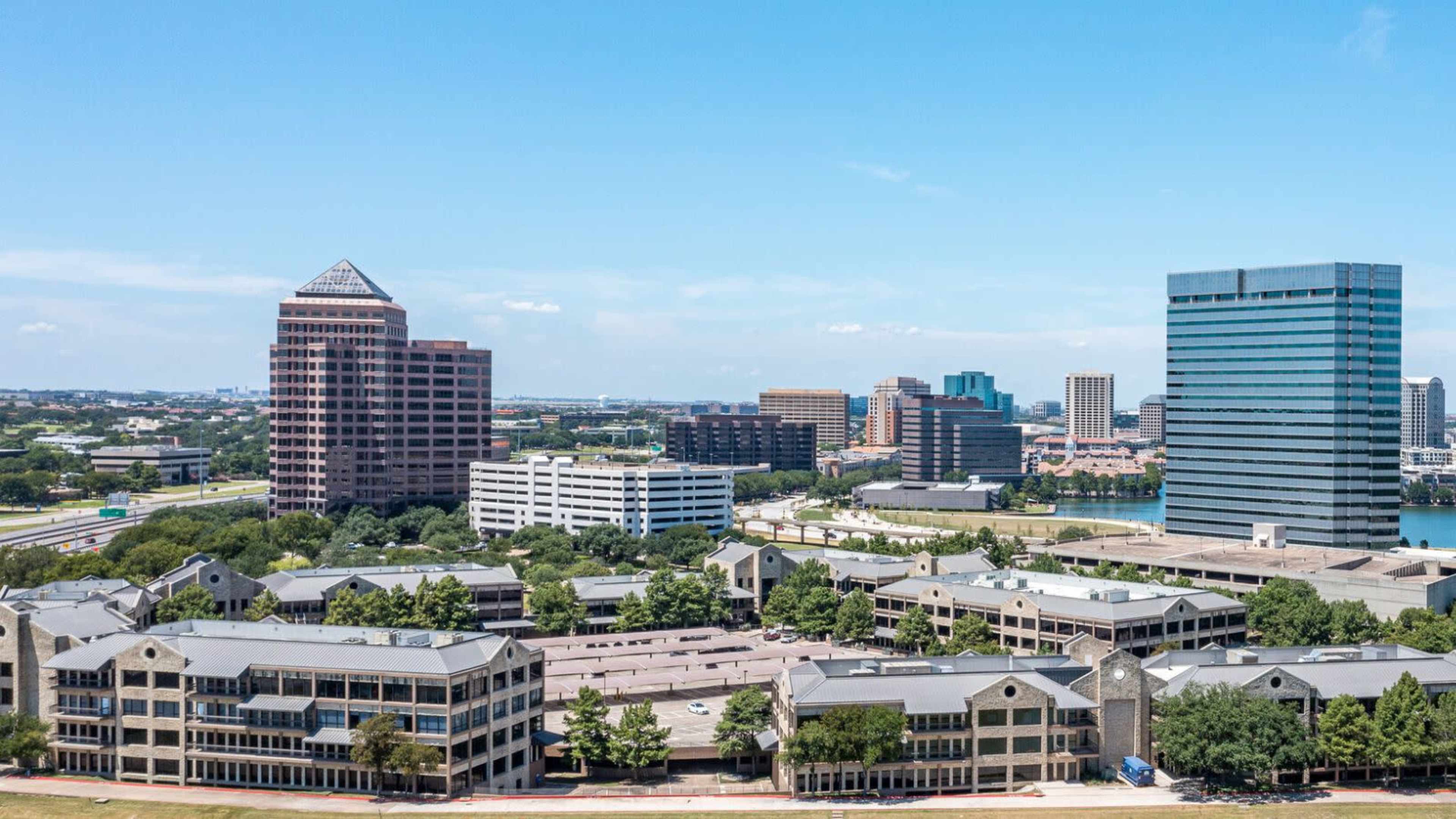 A city skyline featuring a mix of modern office buildings and low-rise structures surrounded by greenery, with a clear blue sky overhead.