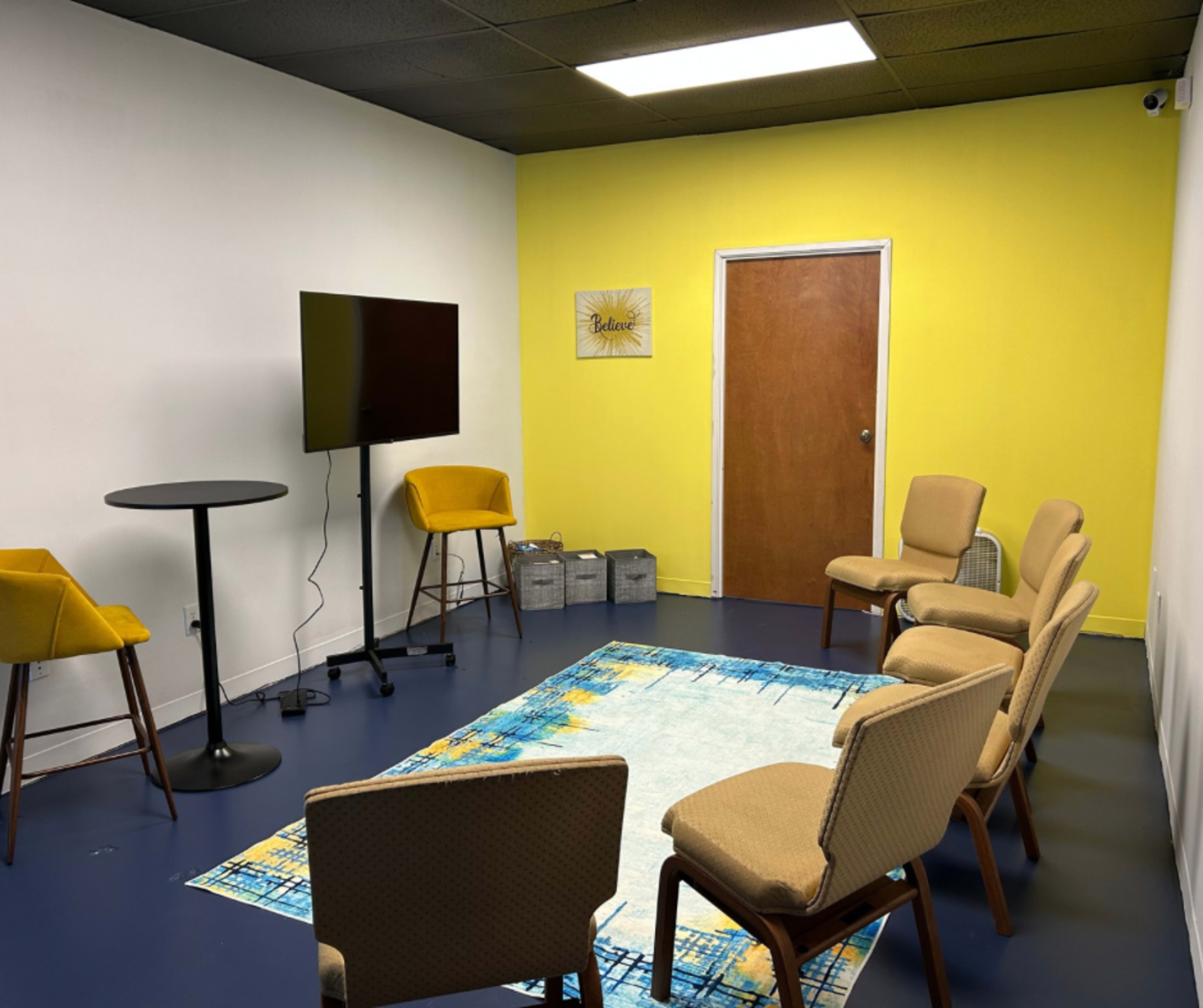 A small meeting room features a round table, yellow walls, a television on a stand, and six chairs arranged around a colorful area rug.