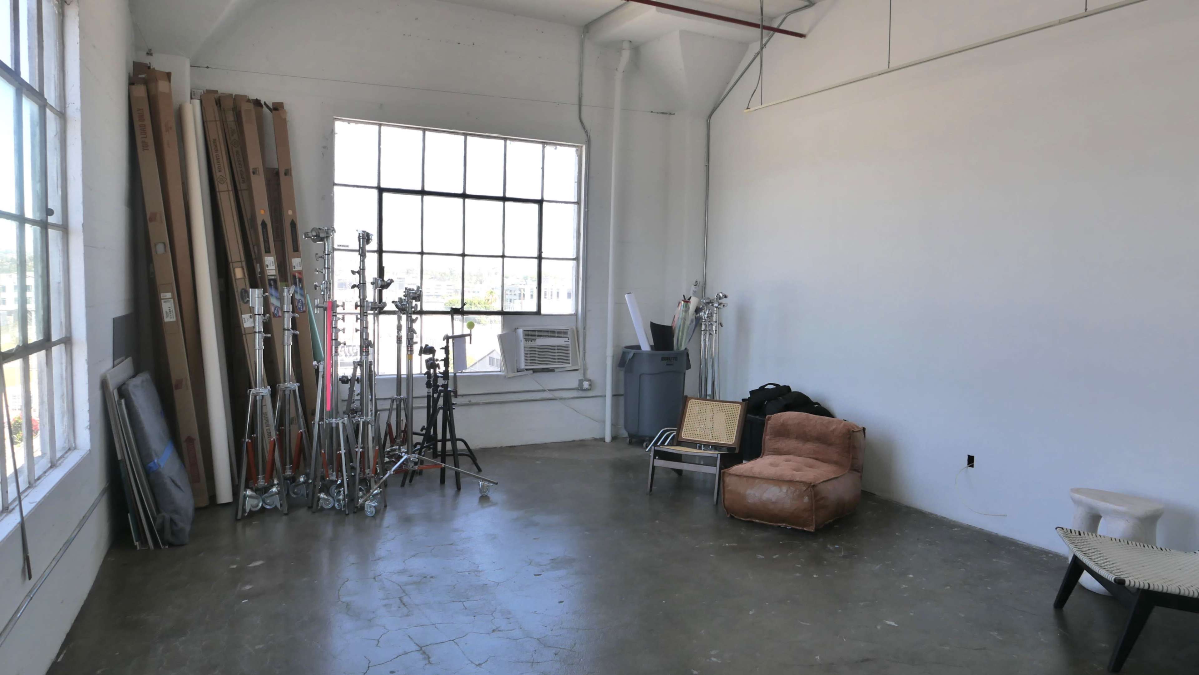 The image shows a spacious, empty studio with large windows, various photography stands stacked against the wall, and a couple of chairs in one corner.