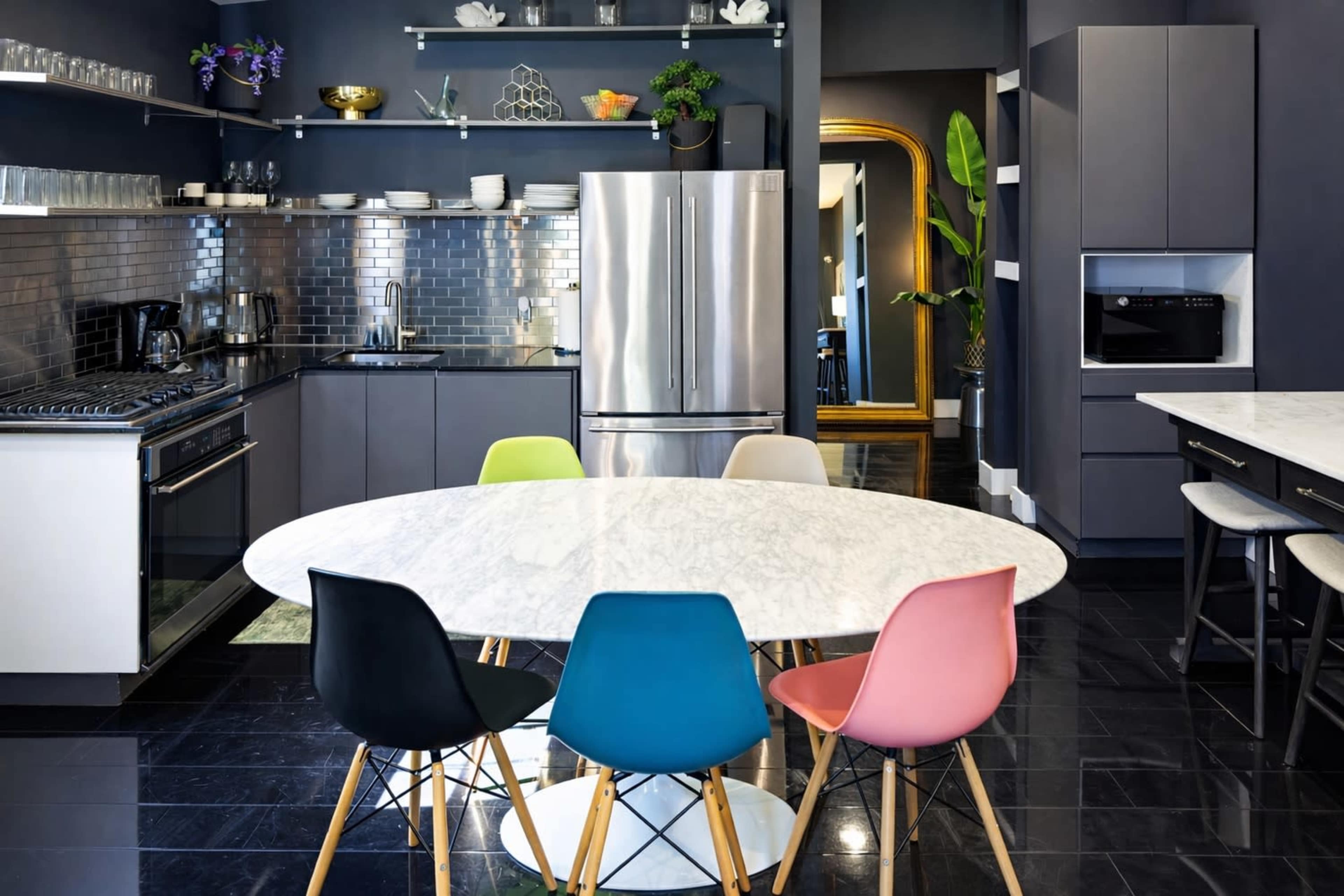 The image shows a modern kitchen with gray cabinetry, a stainless steel refrigerator, and a large marble oval dining table surrounded by colorful chairs.