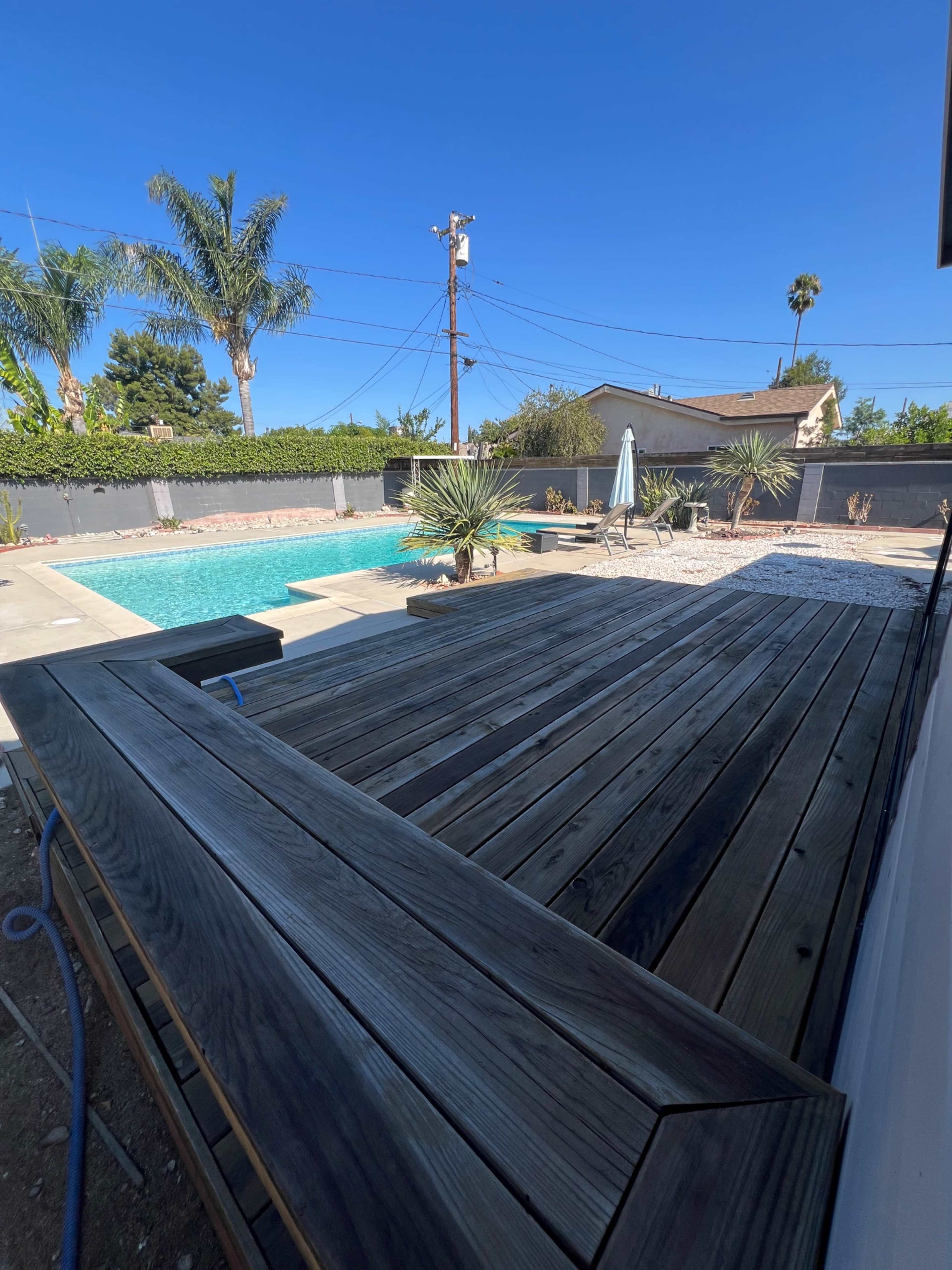 A wooden deck overlooks a swimming pool surrounded by palm trees and desert plants in a residential area.