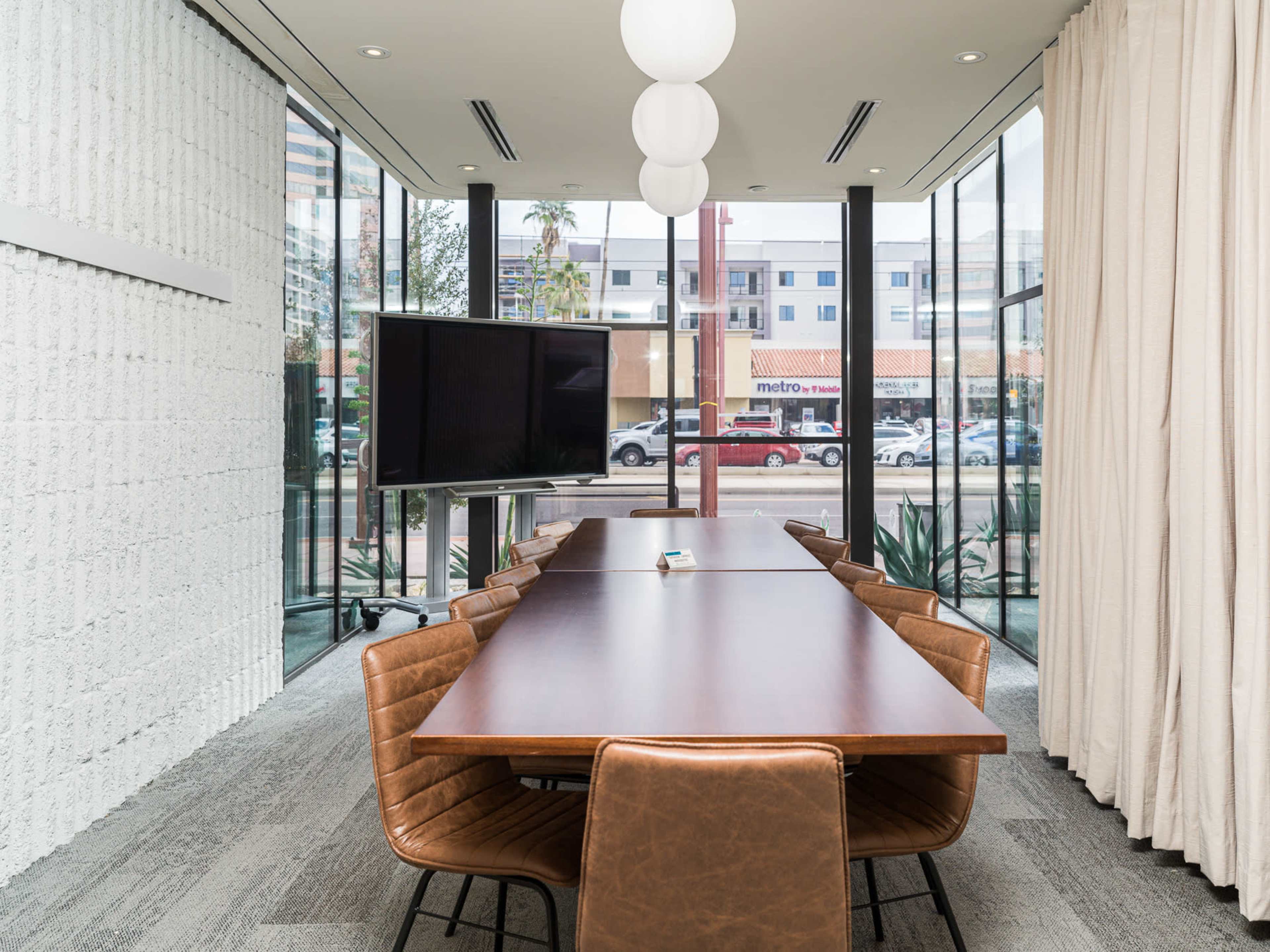 A modern conference room featuring a long wooden table, leather chairs, large windows, and a television screen.
