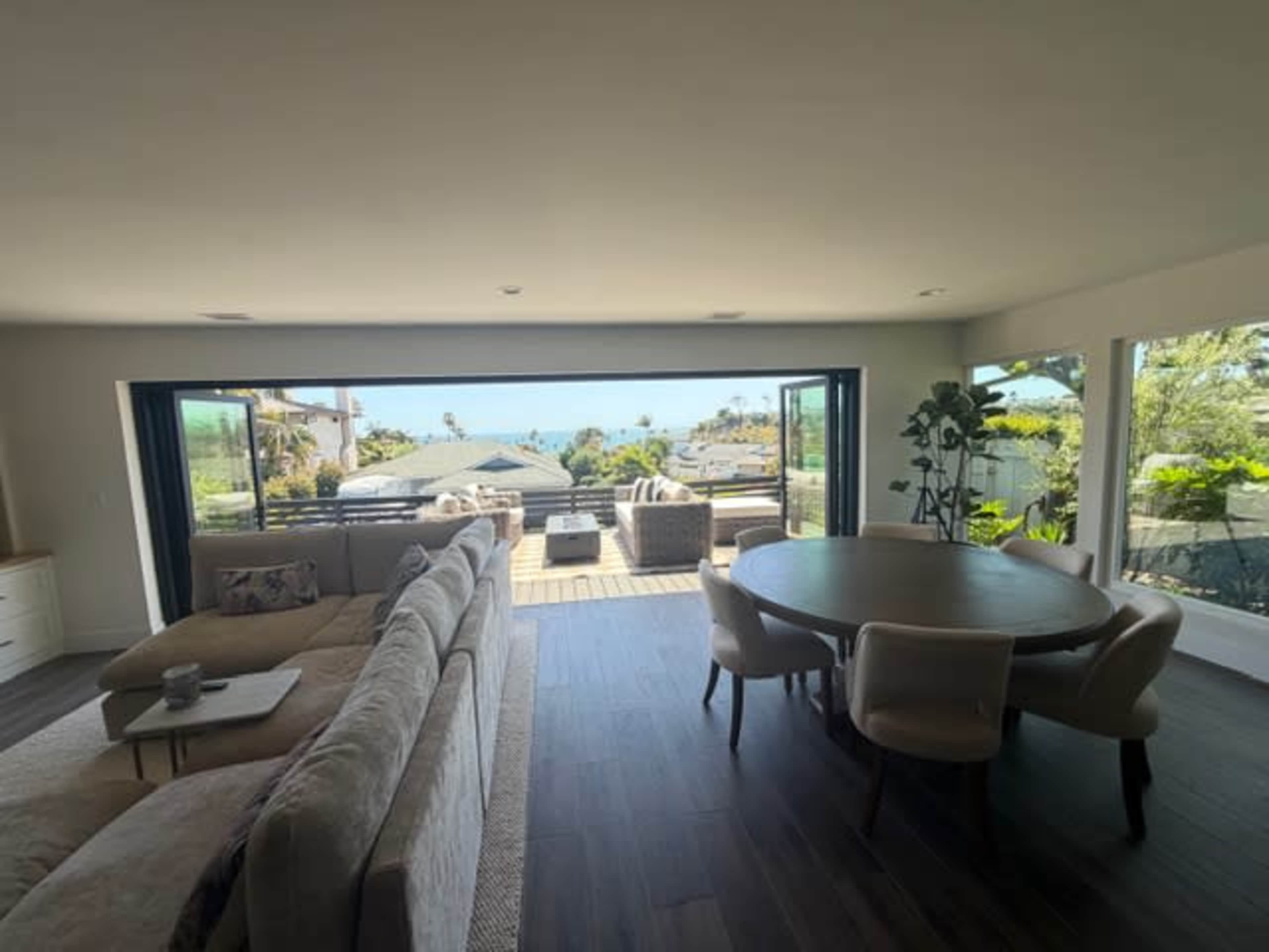 The image shows a modern living room with large sliding glass doors opening to a deck and views of the ocean and surrounding greenery.