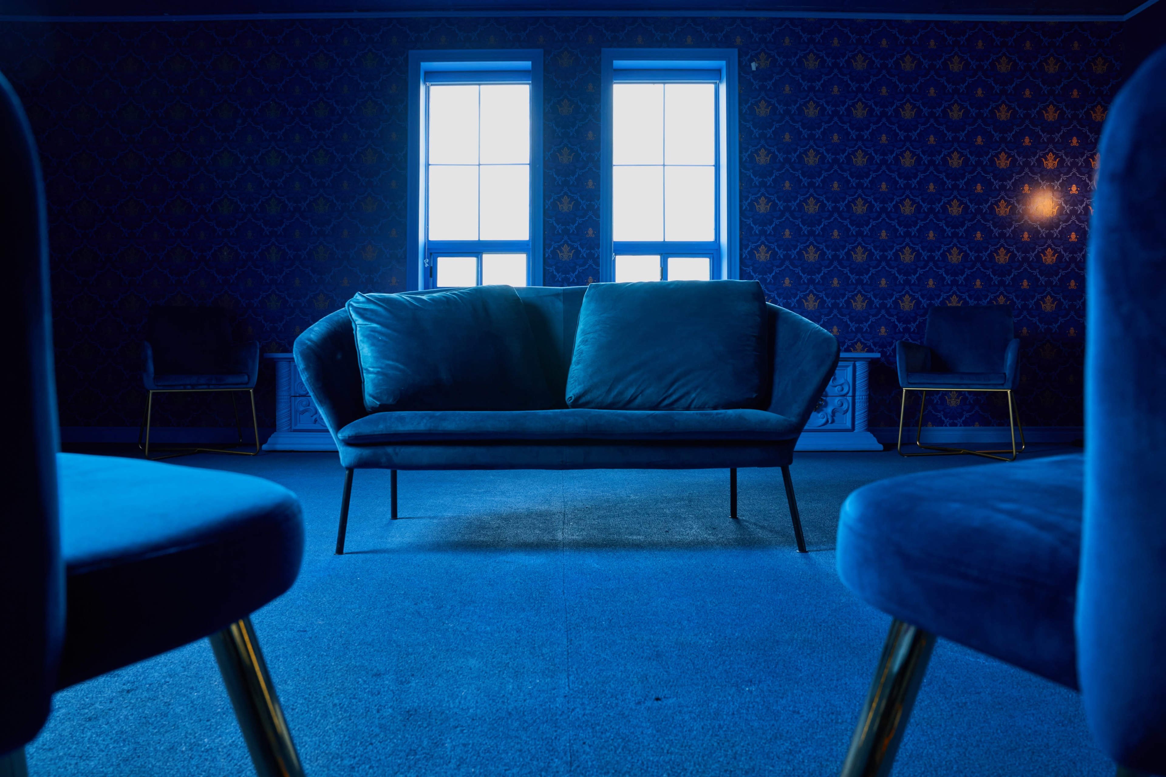 A blue-toned room features a central velvet sofa facing two windows, with two chairs in the background.