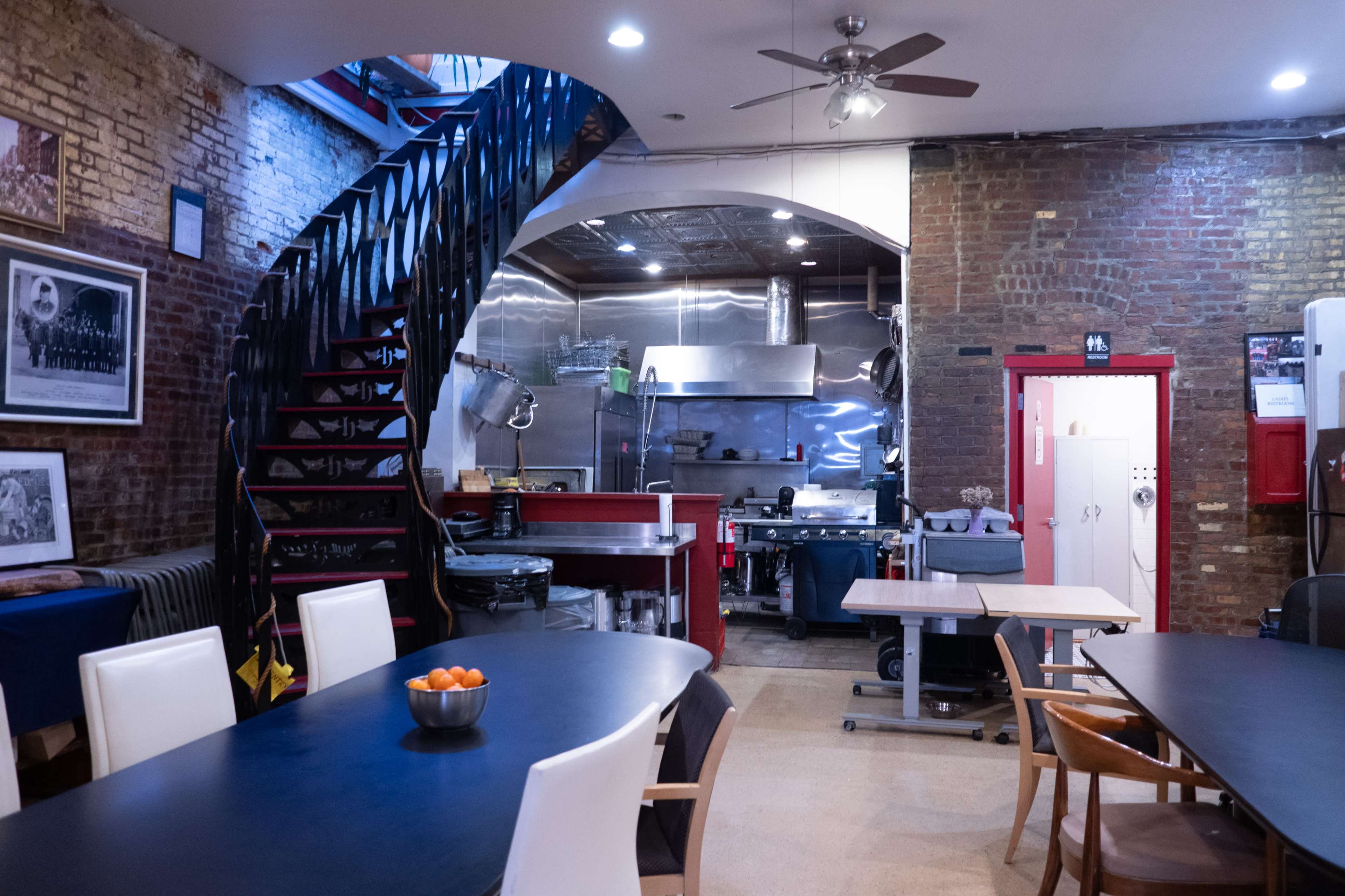The image shows a spacious dining area with a large black table and white chairs, adjacent to an industrial-style kitchen featuring stainless steel appliances and a spiral staircase leading to an upper level.
