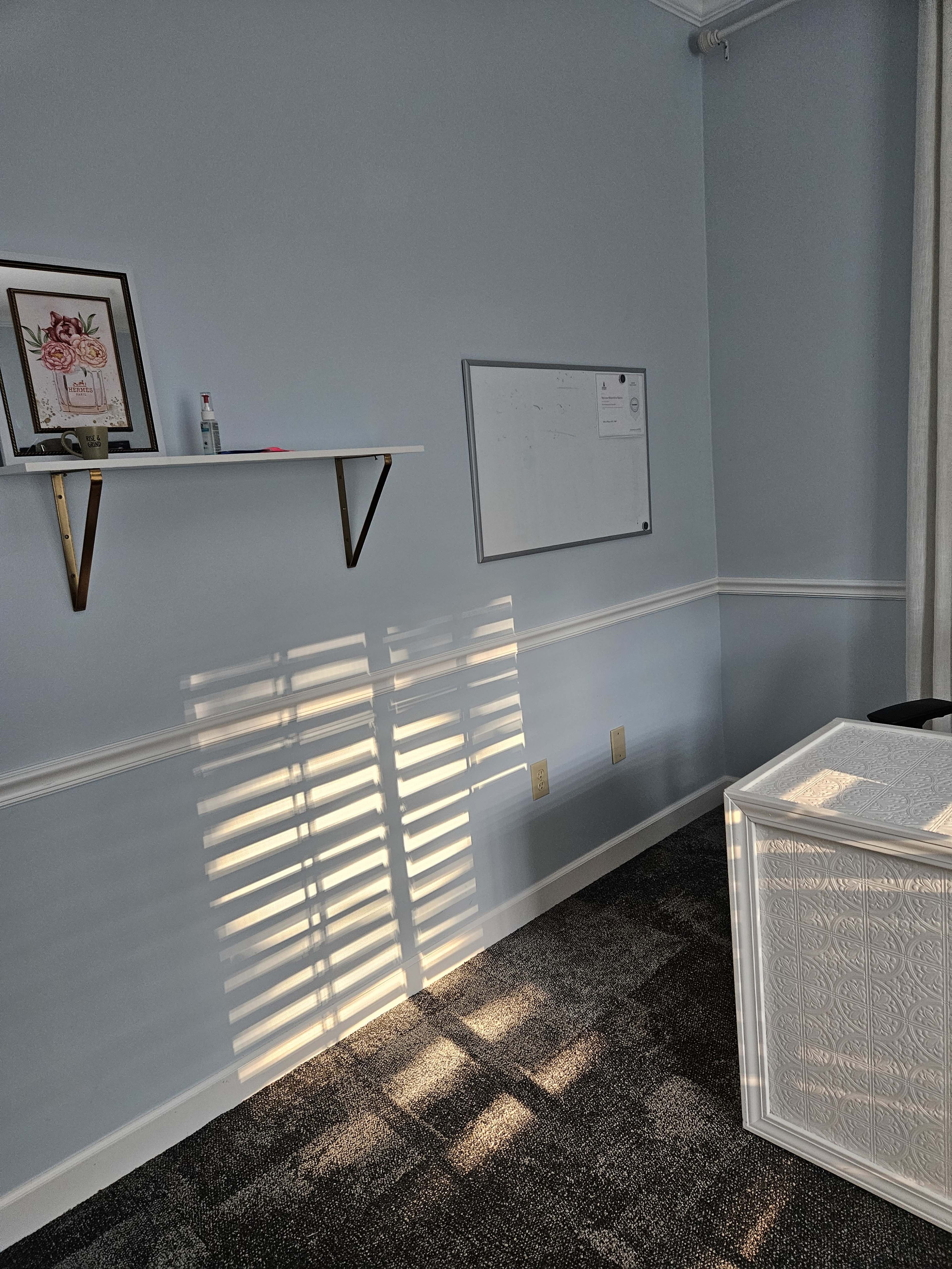 The image shows a bright, empty office space with a whiteboard and a shelf on the wall, along with shadows cast by window blinds on the carpeted floor.
