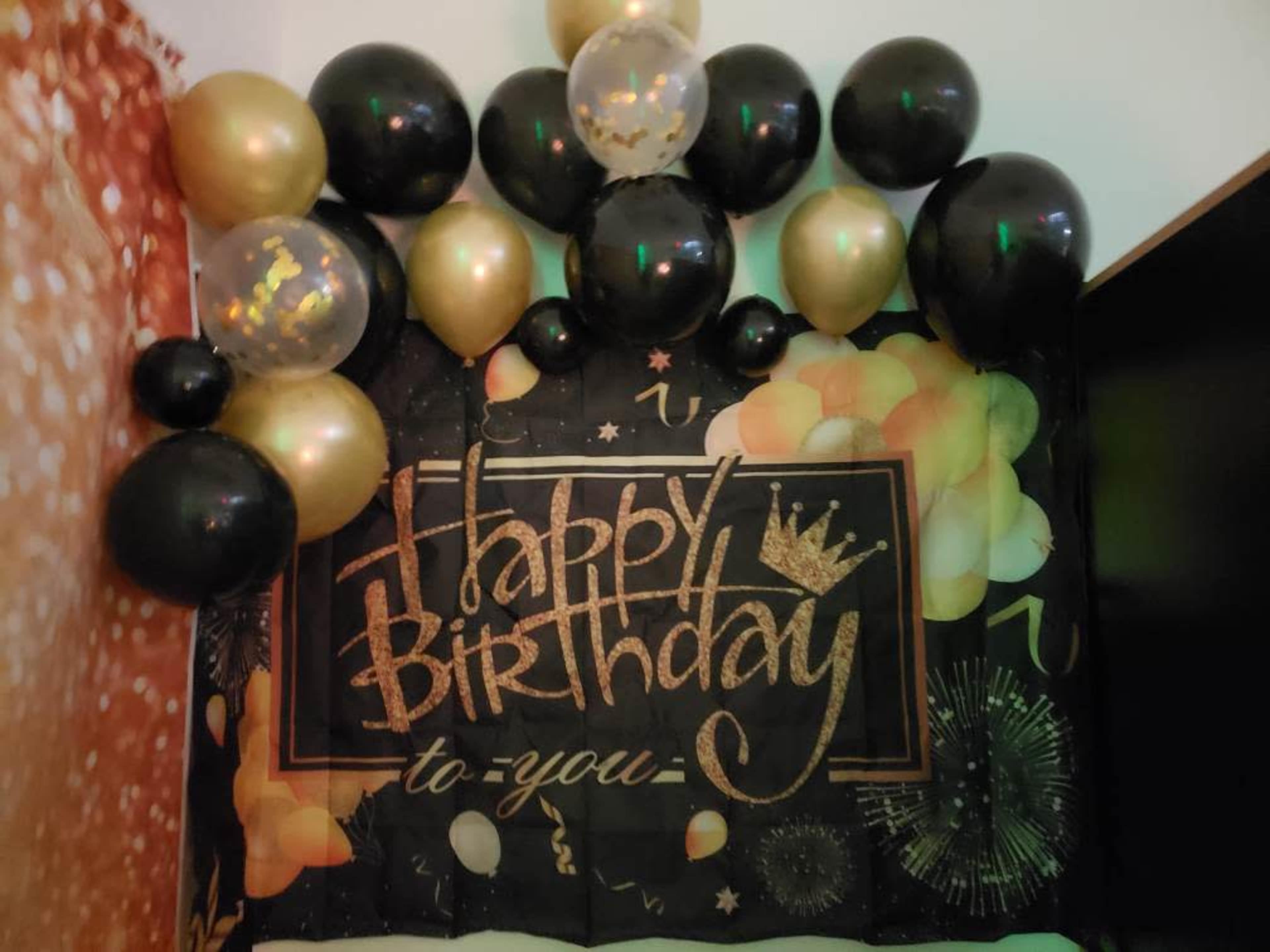A birthday decoration features a black backdrop with "Happy Birthday to You" written in gold, surrounded by black, gold, and transparent balloons.
