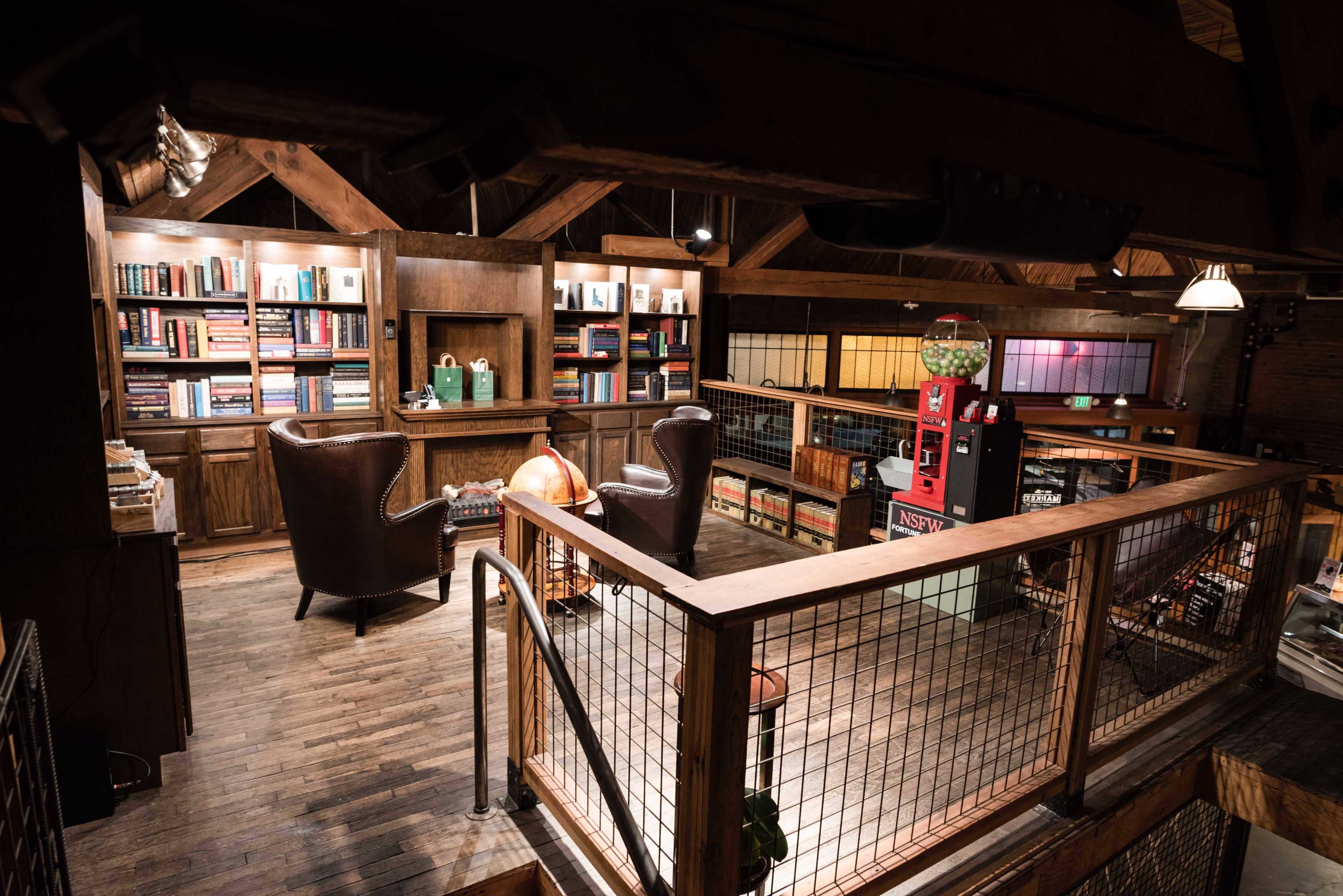 A cozy upper level of a wooden library or reading room with bookshelves, comfortable chairs, and a vintage vending machine.