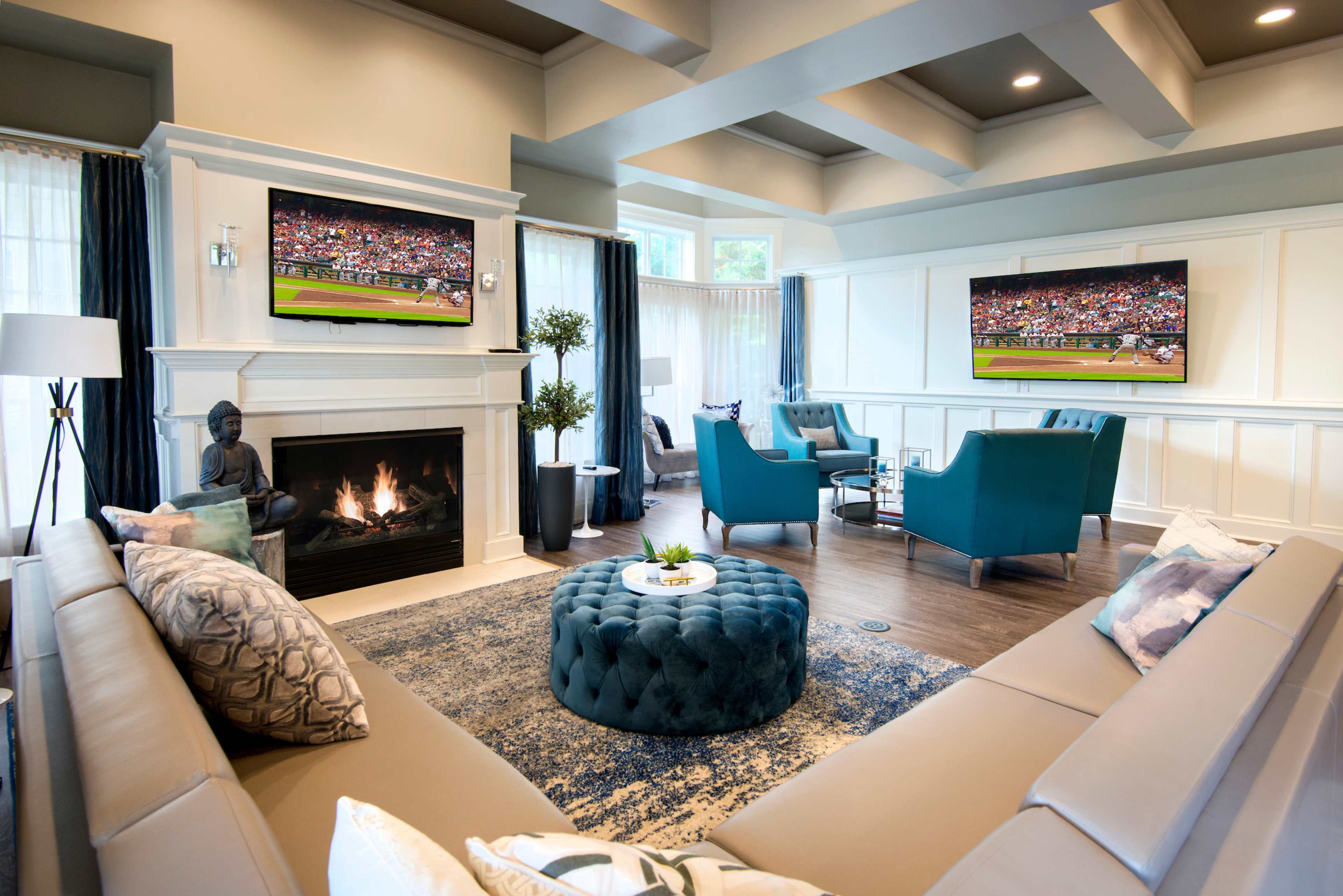 A modern living room with a fireplace, two teal chairs, a large sectional sofa, and two televisions displaying a sports event on opposite walls.