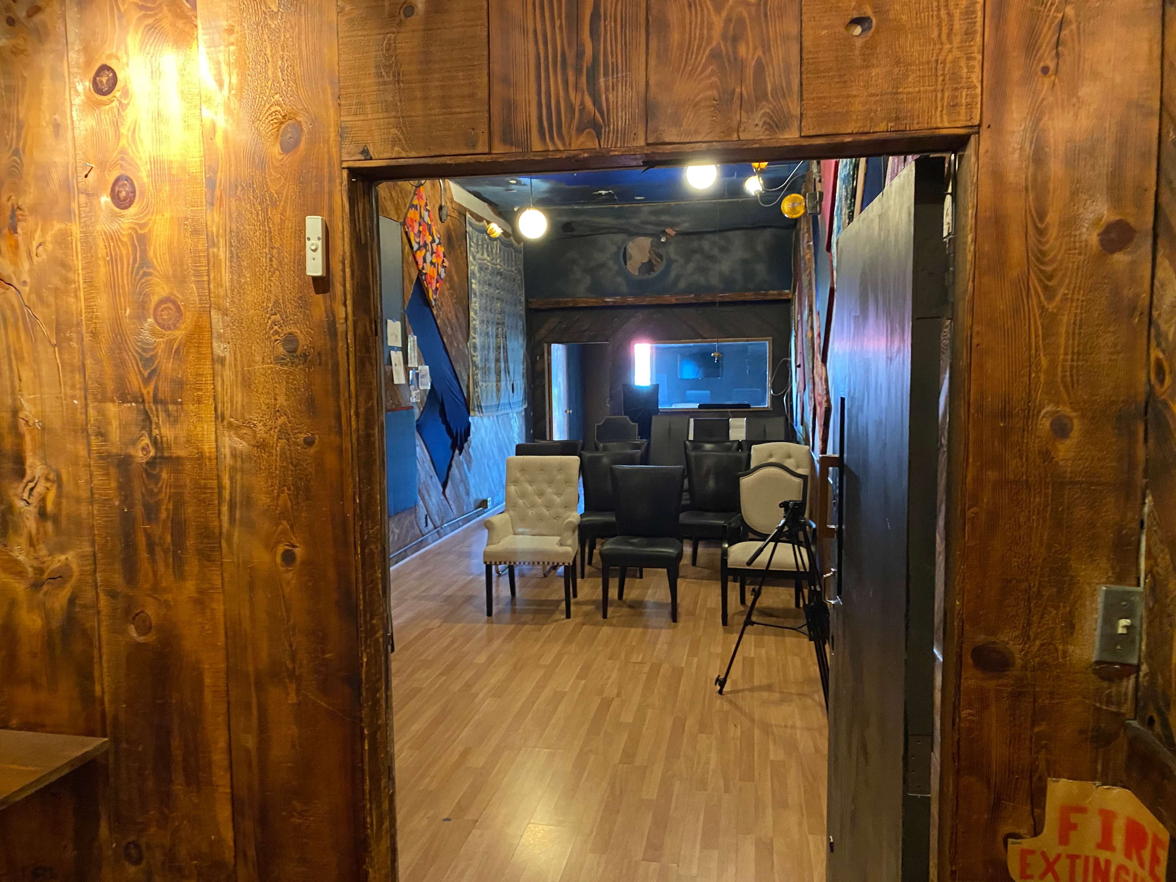 The scene shows a wooden doorway leading to a room with several chairs arranged in rows and a tripod set up in the back.