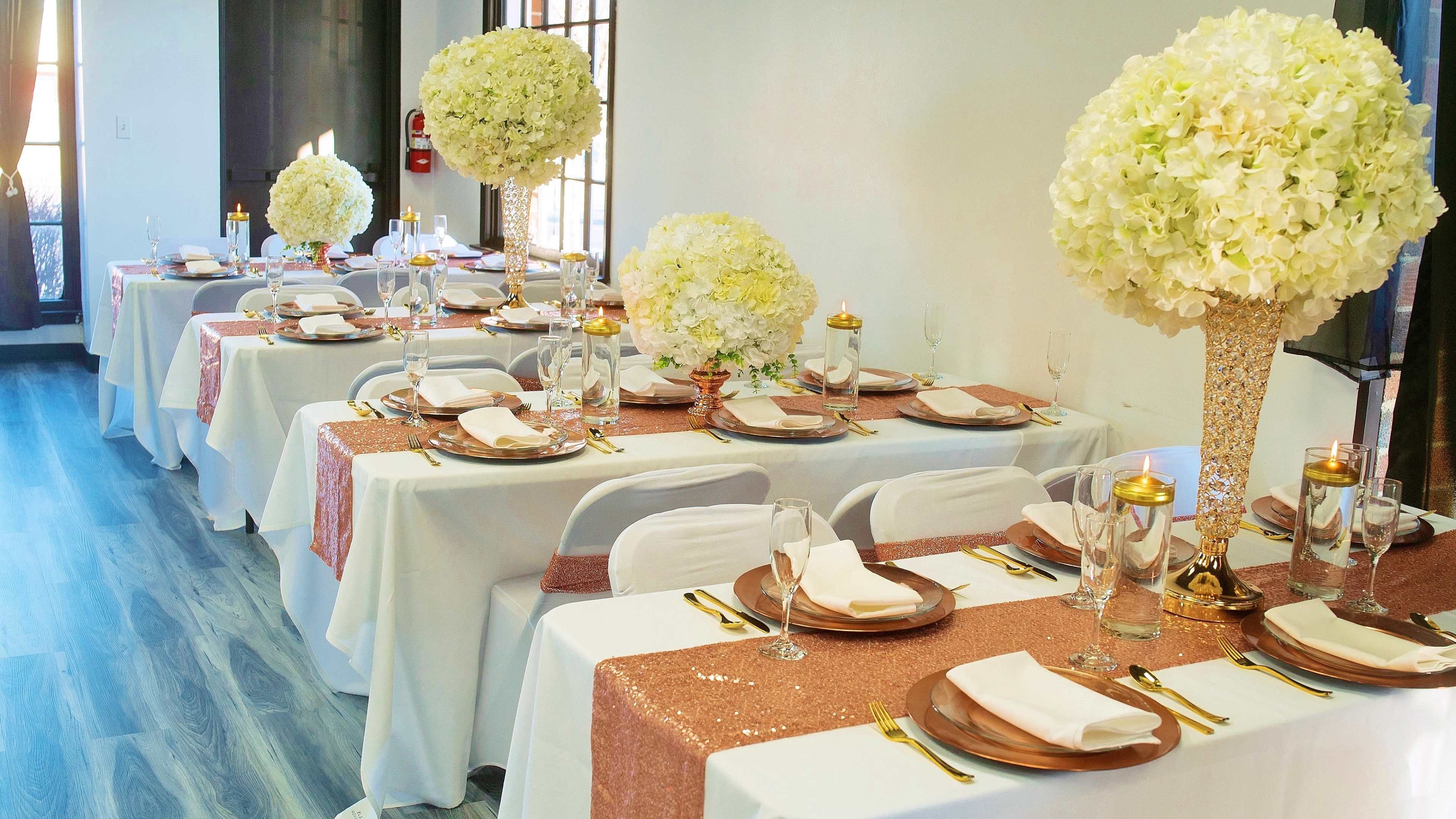 A dining setup featuring multiple tables dressed in white tablecloths with gold and rose-gold accents, adorned with large floral centerpieces and elegant tableware.