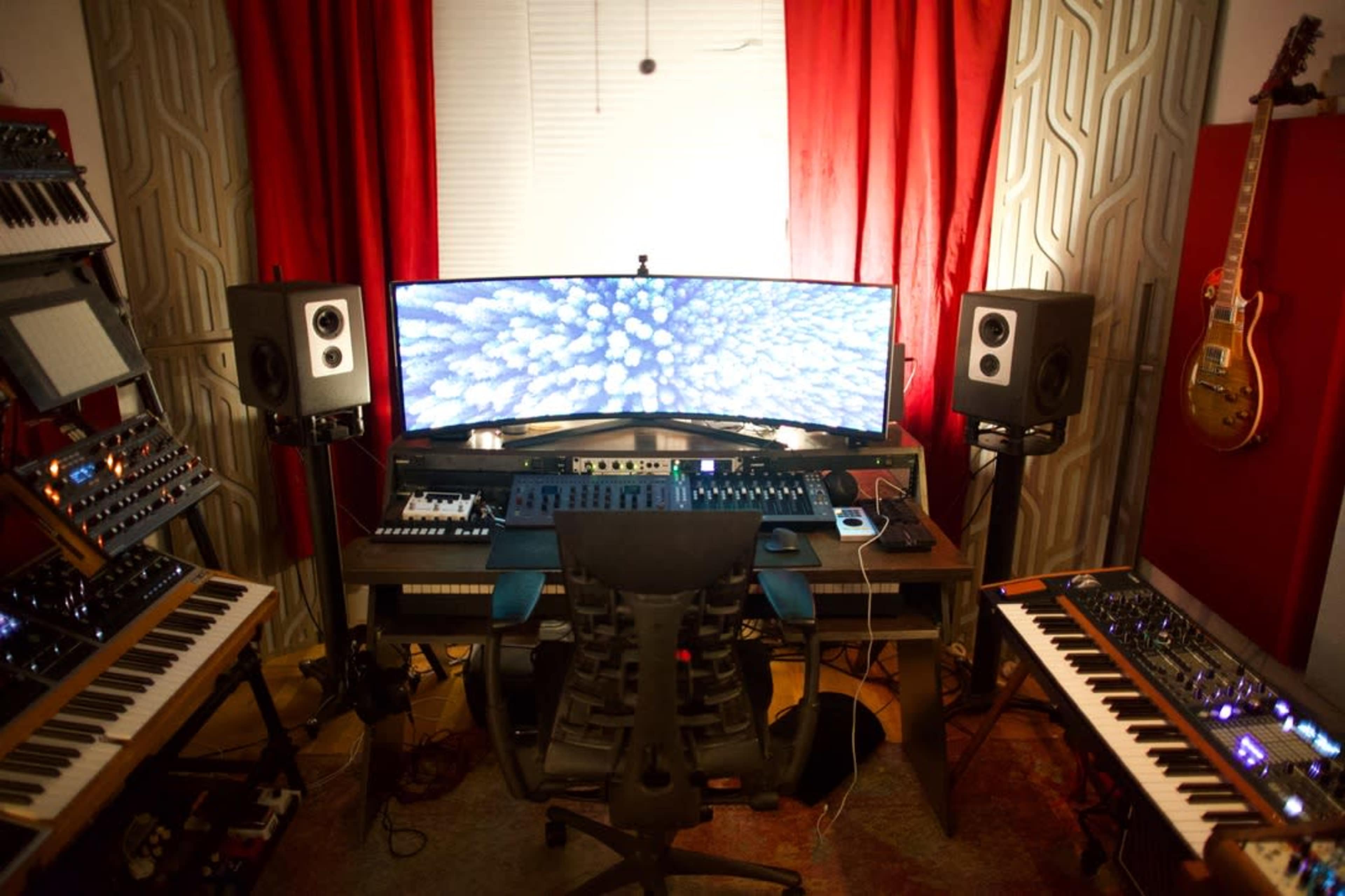 A music production studio features a curved monitor, synthesizers, speakers, and a chair positioned at a central workstation surrounded by soundproofing panels and a guitar.