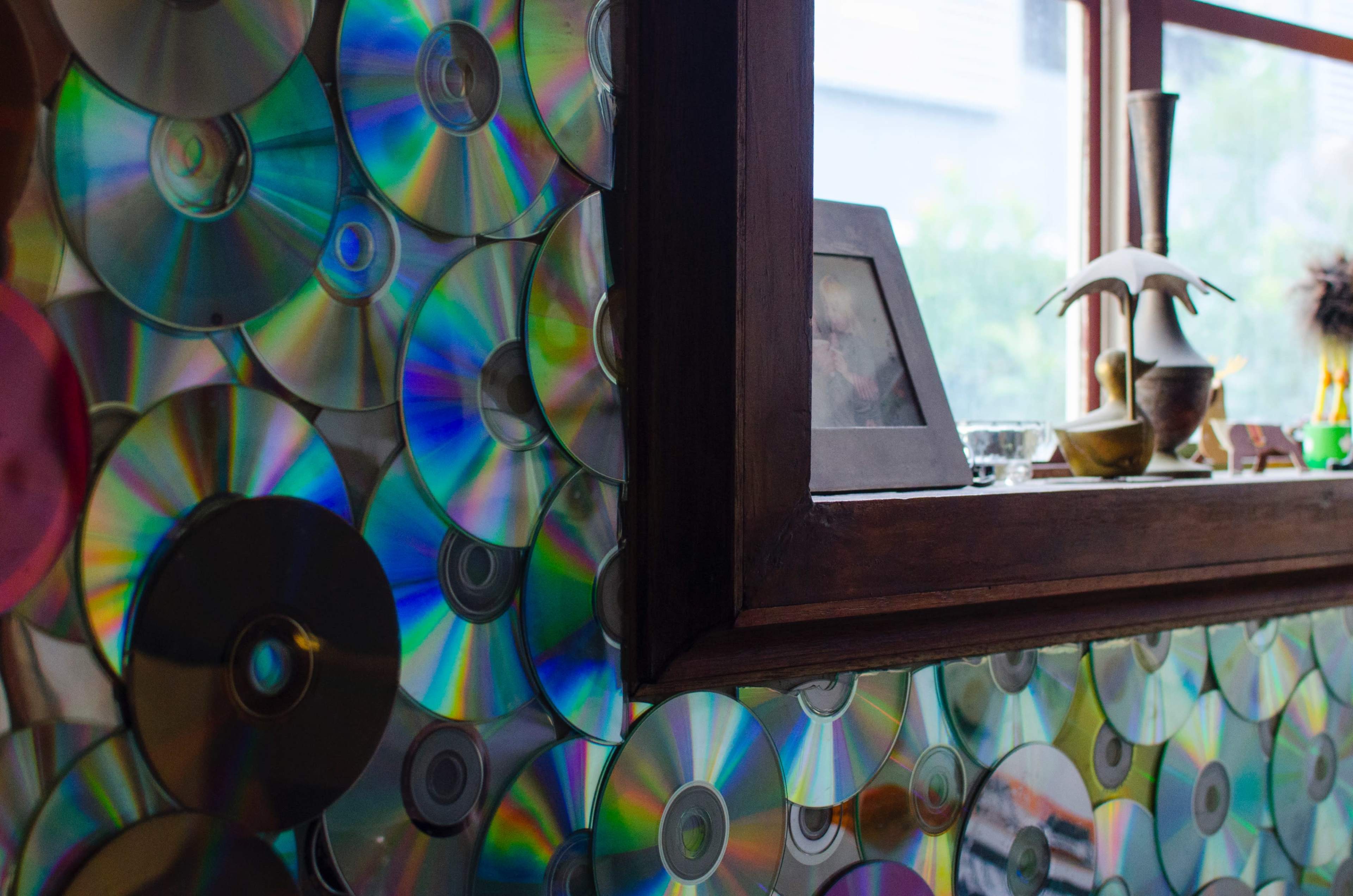 The image shows a wall covered with shiny, colorful CDs and a wooden window frame displaying decorative items.