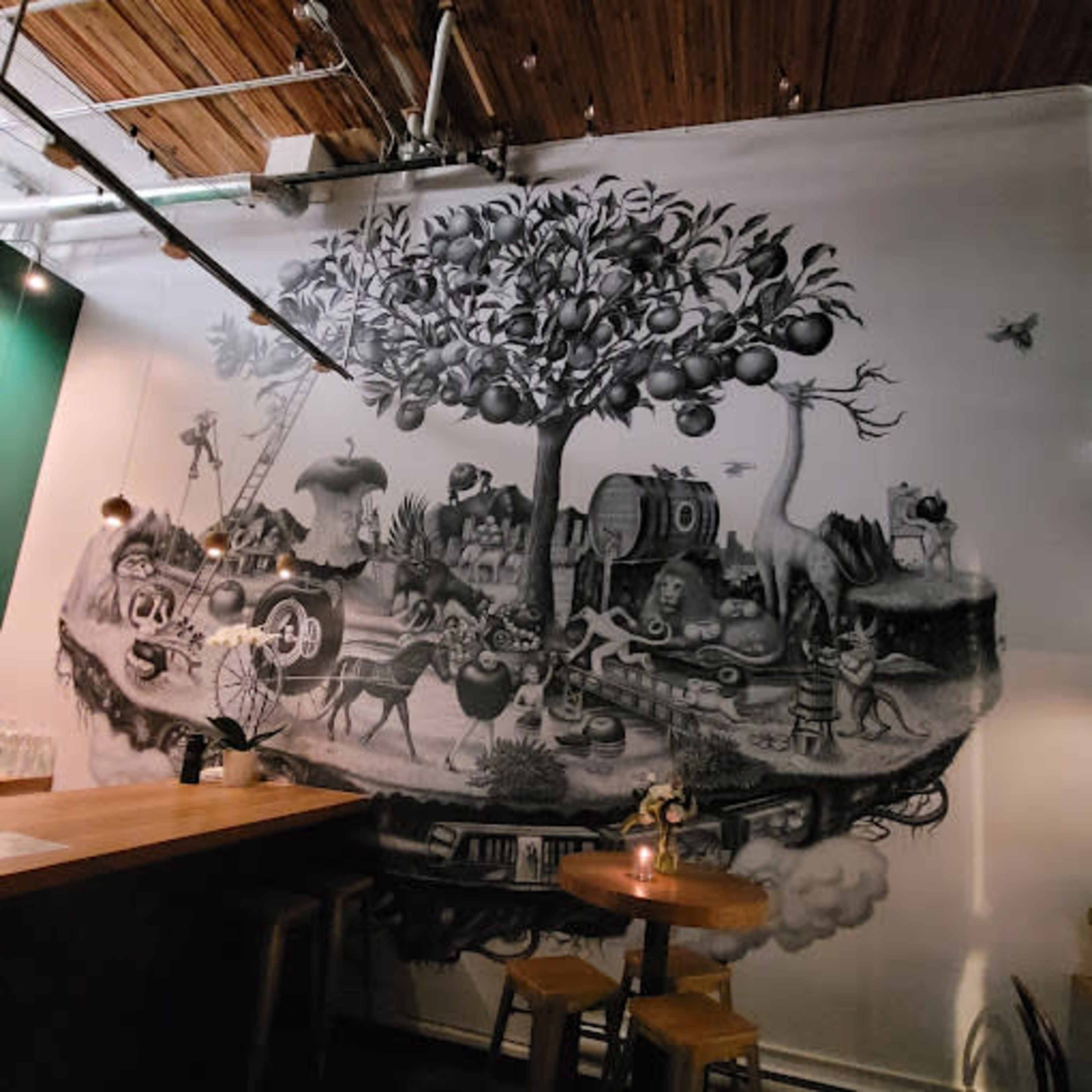 The image shows a large black-and-white mural depicting a whimsical landscape with various characters, animals, and objects, all interconnected beneath a tree with fruit.