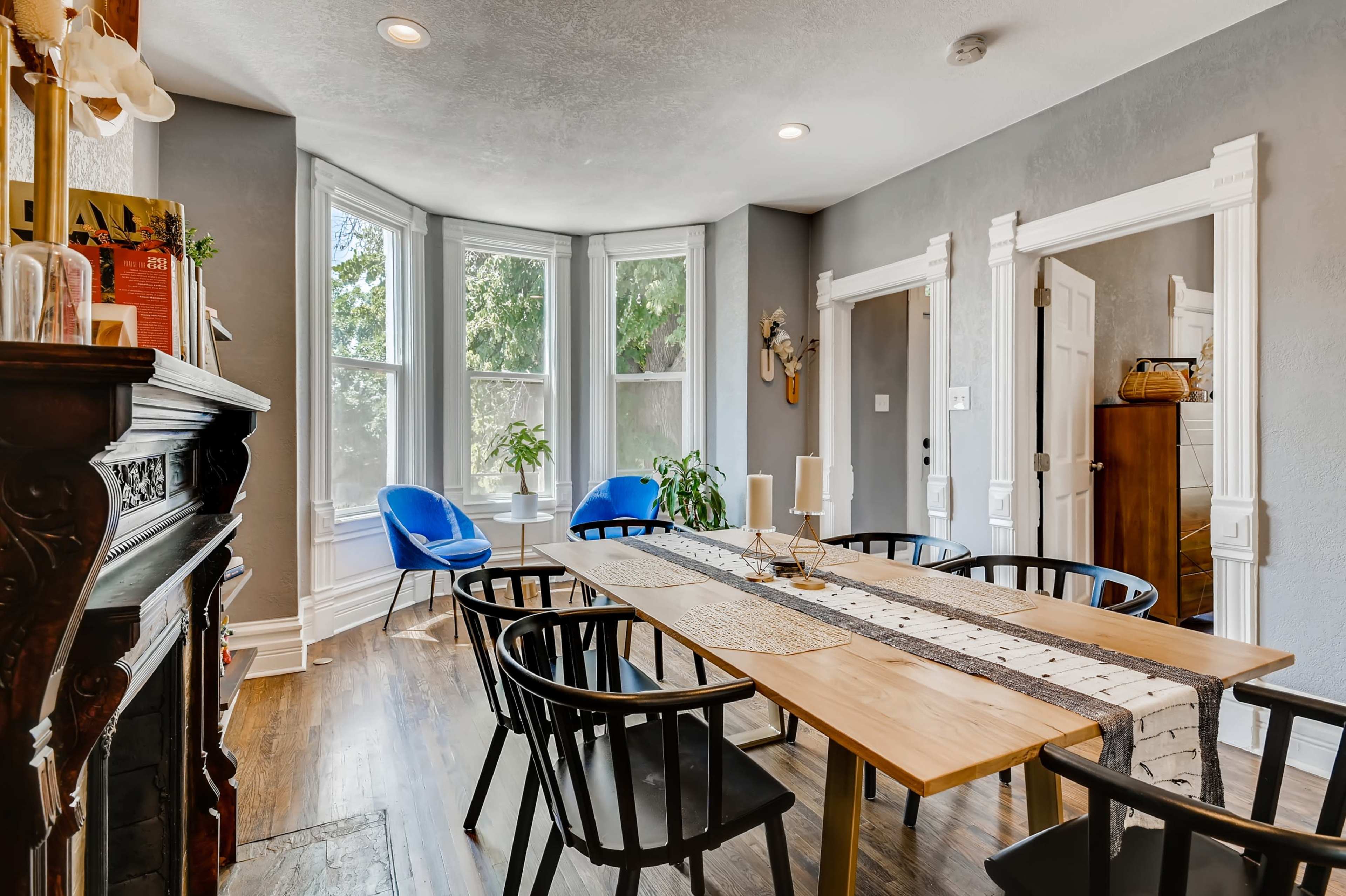 A dining room features a long wooden table surrounded by black chairs, with two blue chairs and large windows letting in natural light.