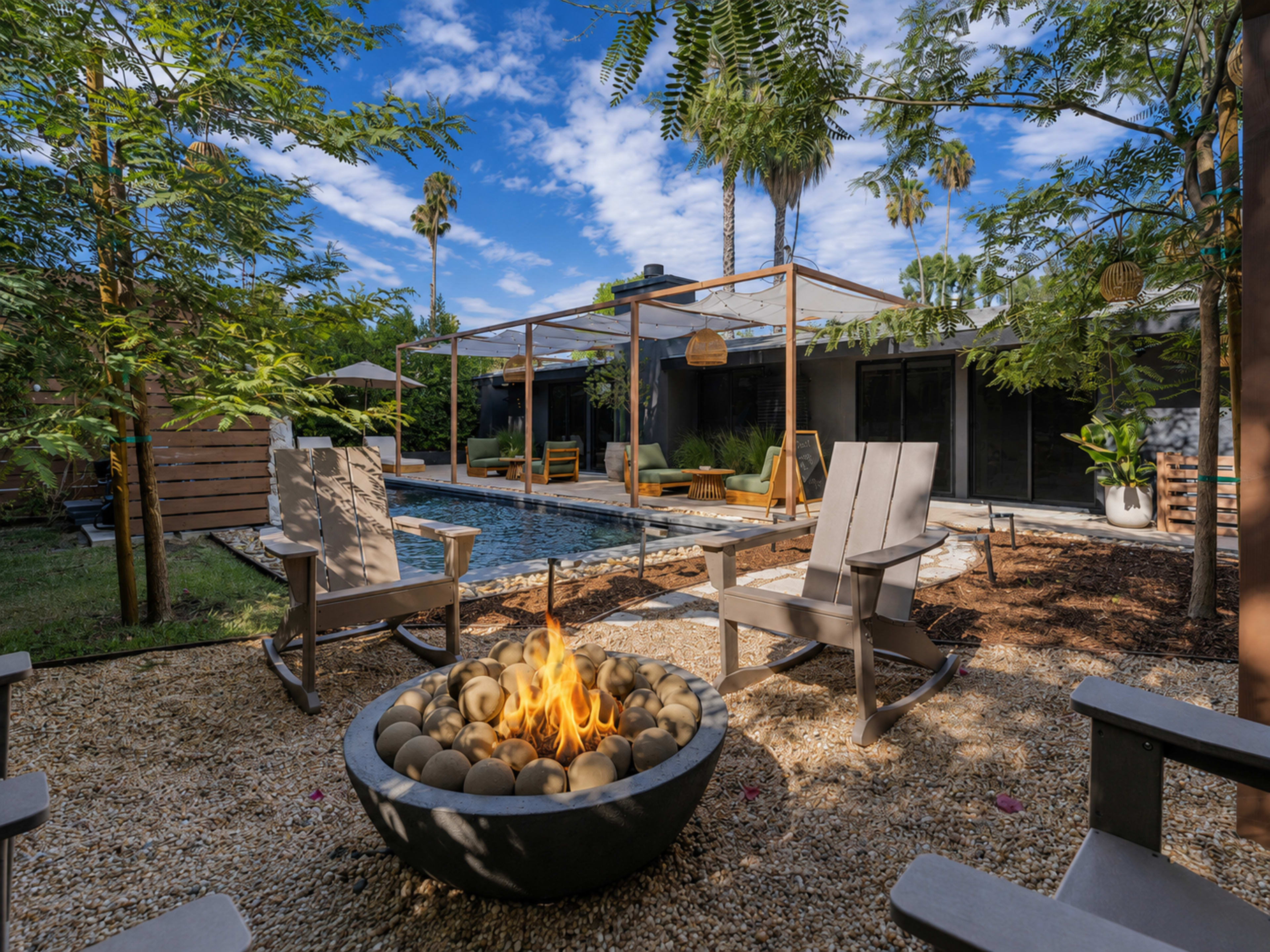 A modern backyard features a fire pit surrounded by chairs, a swimming pool, and palm trees under a clear blue sky.