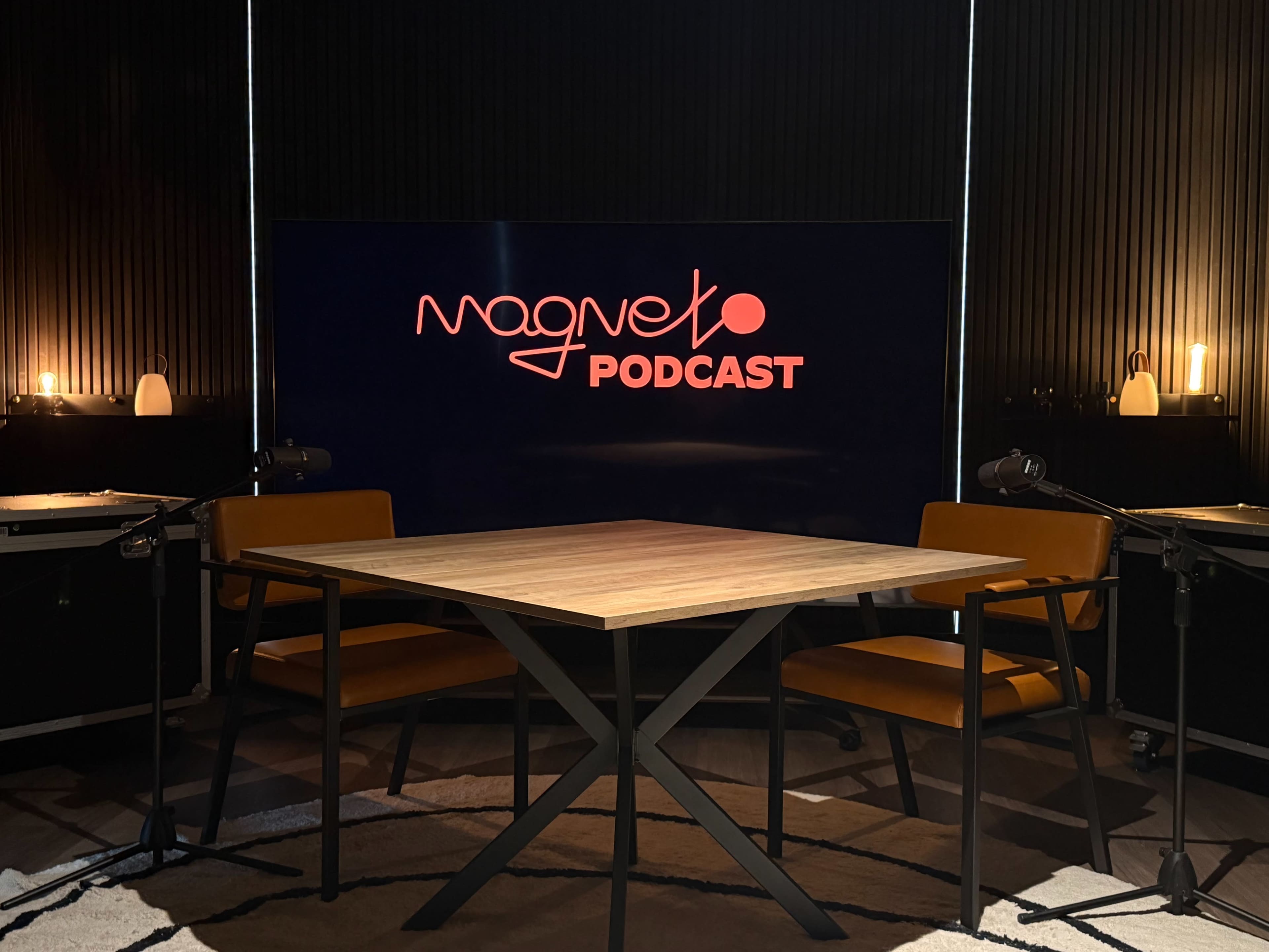 The image shows a podcast recording setup featuring a wooden table and two orange chairs in front of a large screen displaying the text "Magneto Podcast."