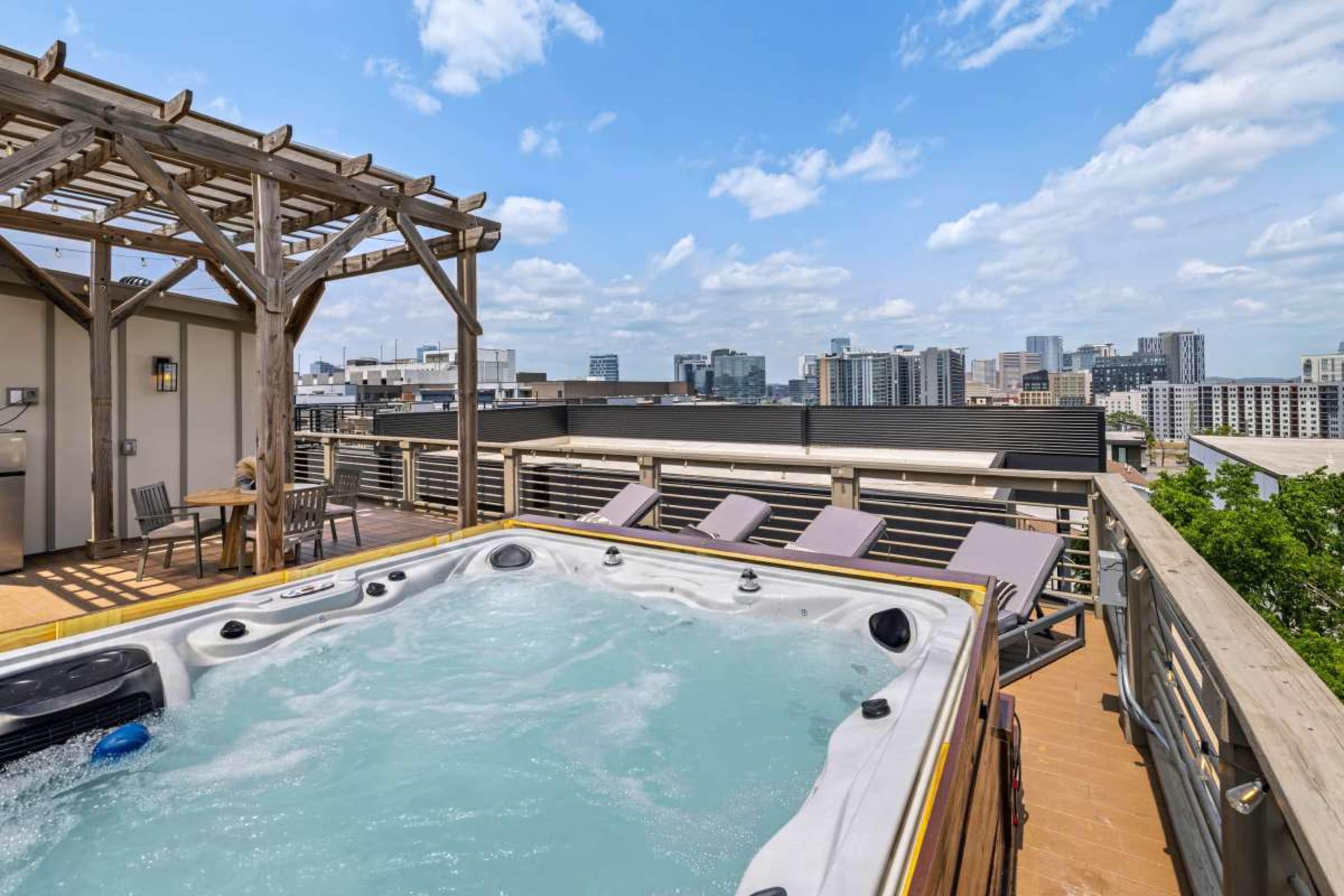 The image shows a rooftop deck with a hot tub overlooking a city skyline, complete with a pergola and lounge chairs.