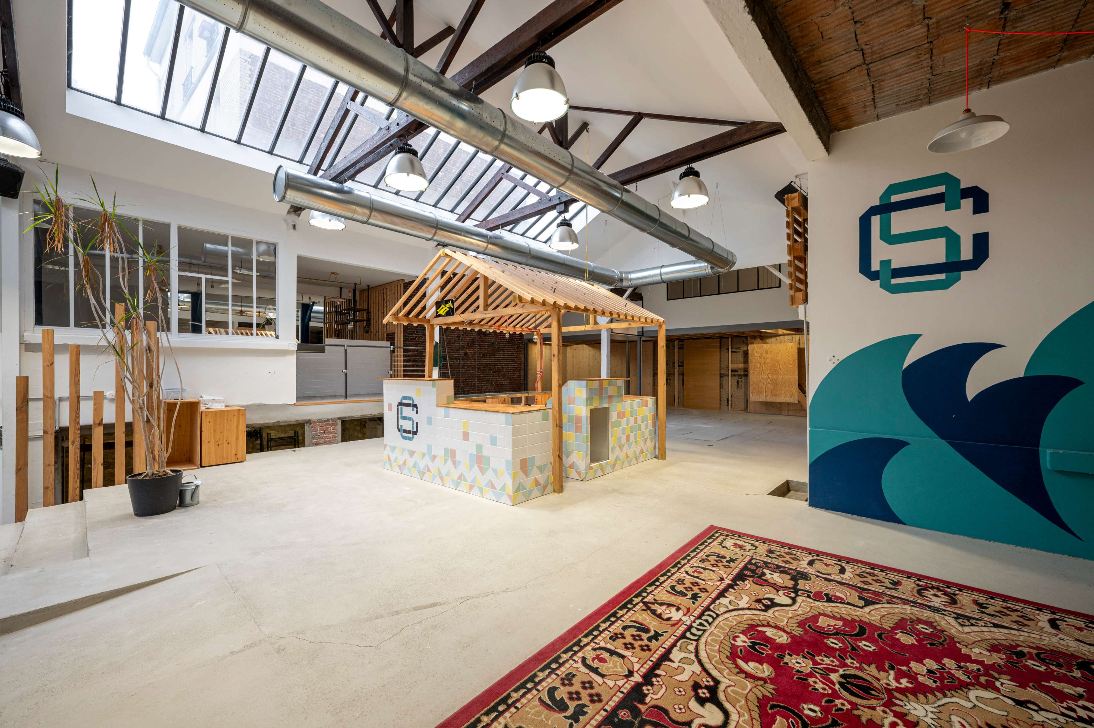 An open workspace featuring a wooden structure resembling a small kiosk, with decorative tiles, a large window above, and a patterned rug on the floor.