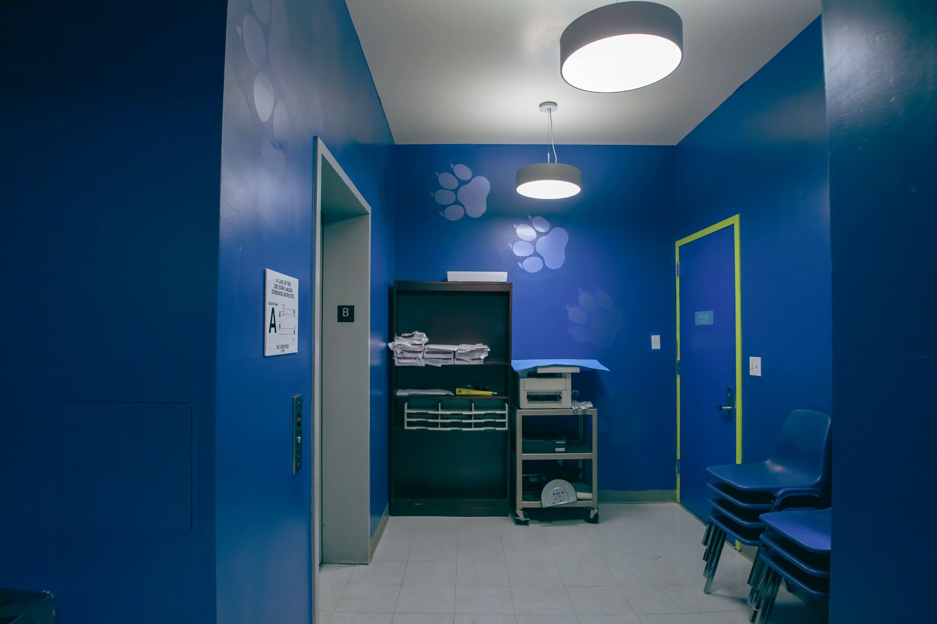 A small, brightly colored room with blue walls, a shelf containing folded towels, a cart with supplies, and stacked chairs in one corner.