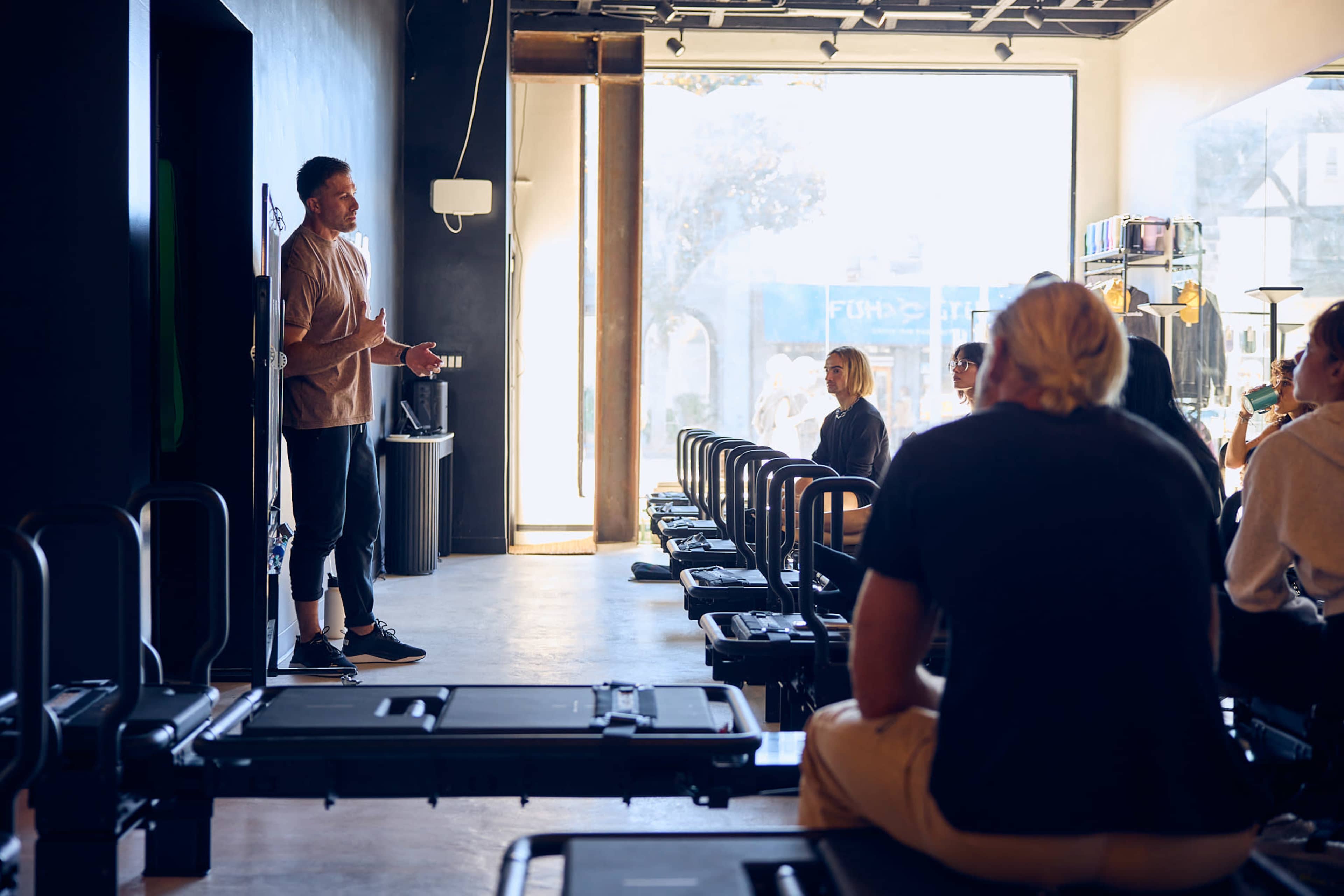 A fitness instructor stands in front of a group of seated individuals in a gym filled with reformer Pilates equipment.