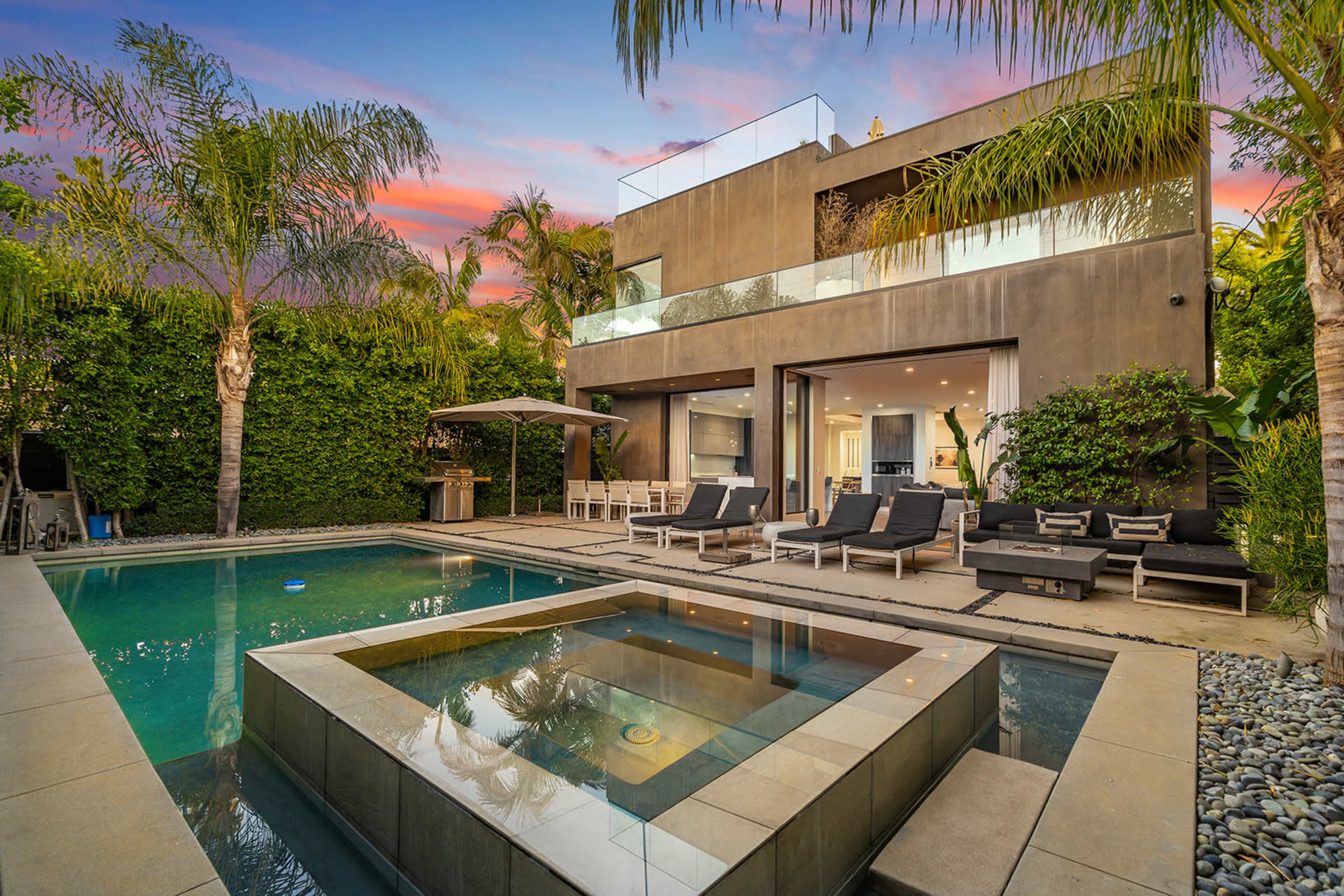 The image shows a modern home with a swimming pool and spa area, surrounded by palm trees and lush greenery, under a colorful sunset sky.