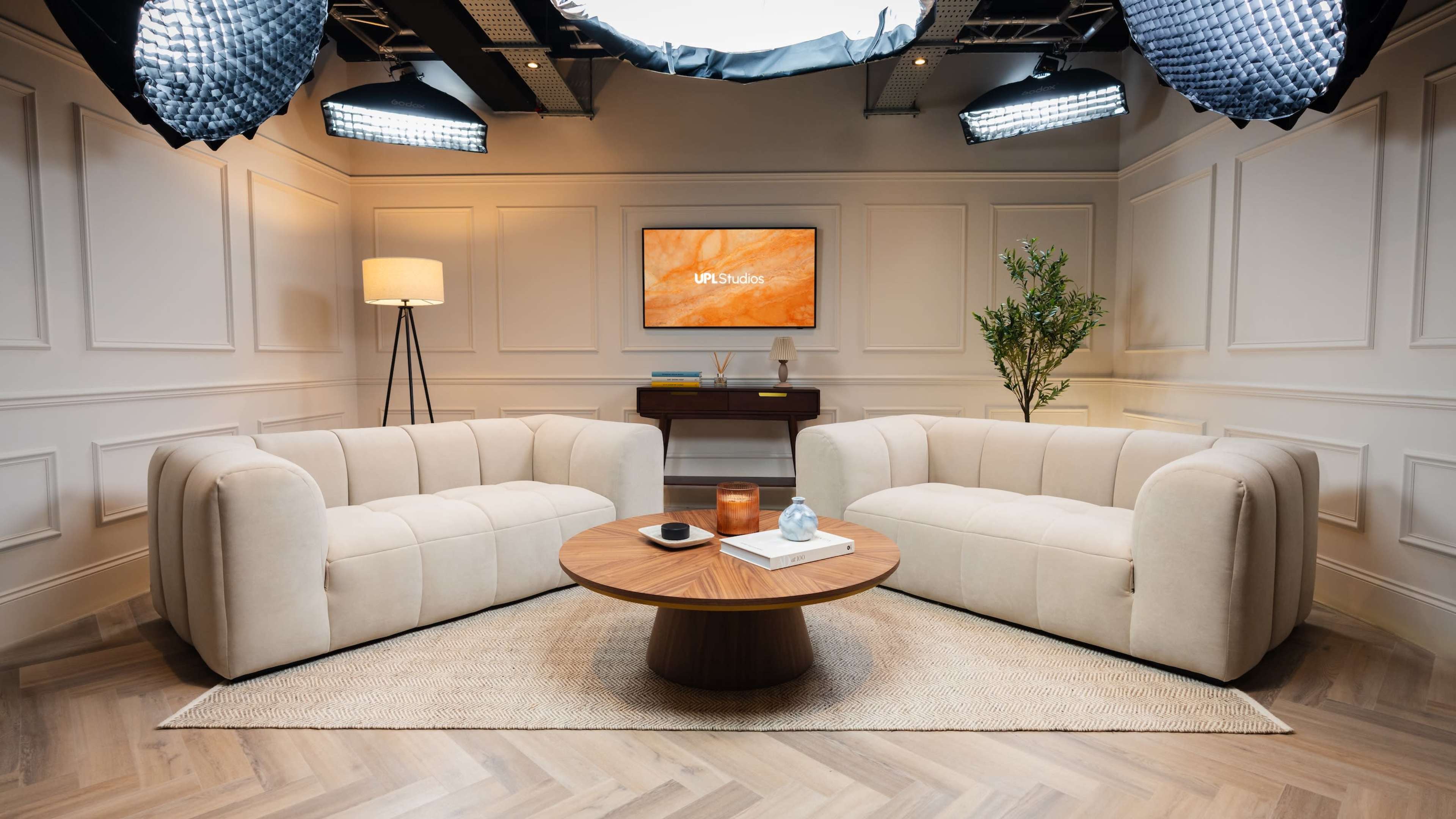 The image showcases a modern studio set featuring two light-colored sofas, a round wooden coffee table, and ambient lighting, with a painting and a plant in the background.
