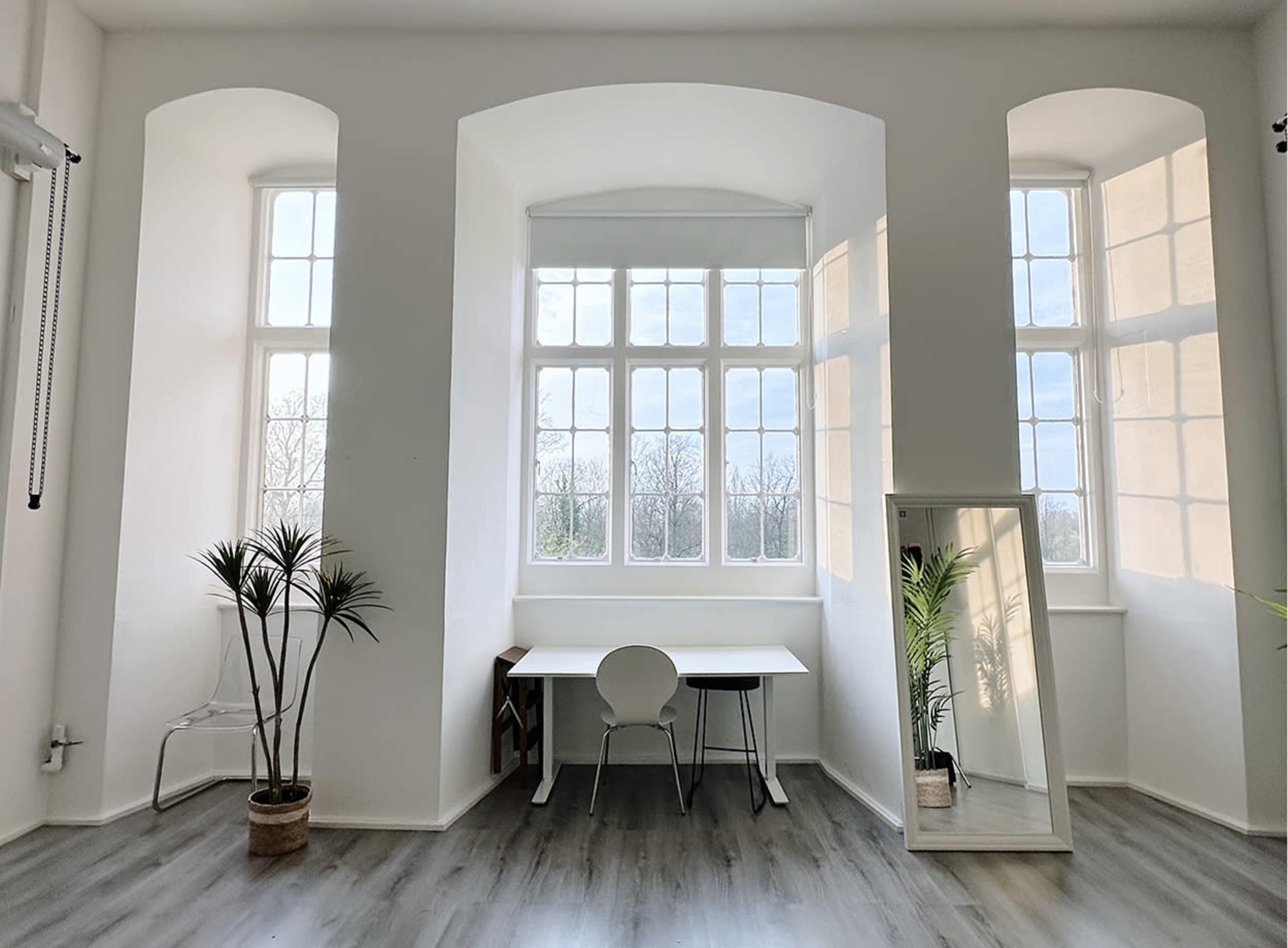 The image depicts a bright, minimalistic workspace featuring large windows, a white desk with a chair, and a mirror beside a potted plant.