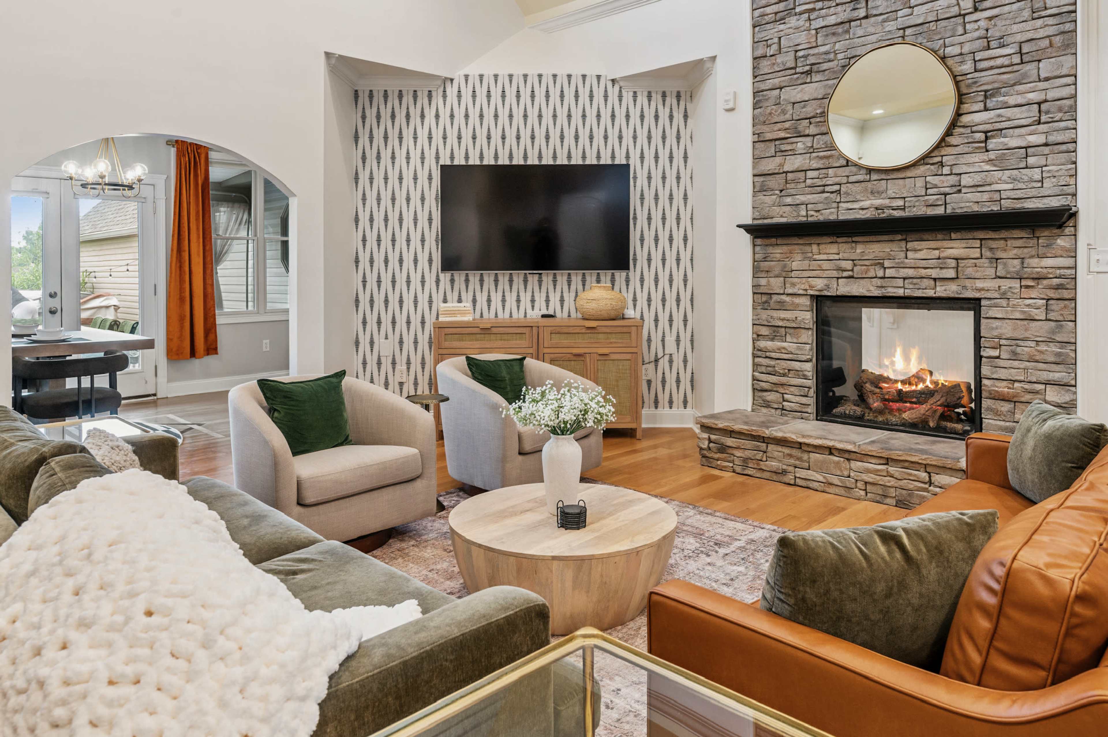 The image shows a cozy living room featuring a stone fireplace, a round wooden coffee table, and a television mounted on the wall, surrounded by comfortable seating.