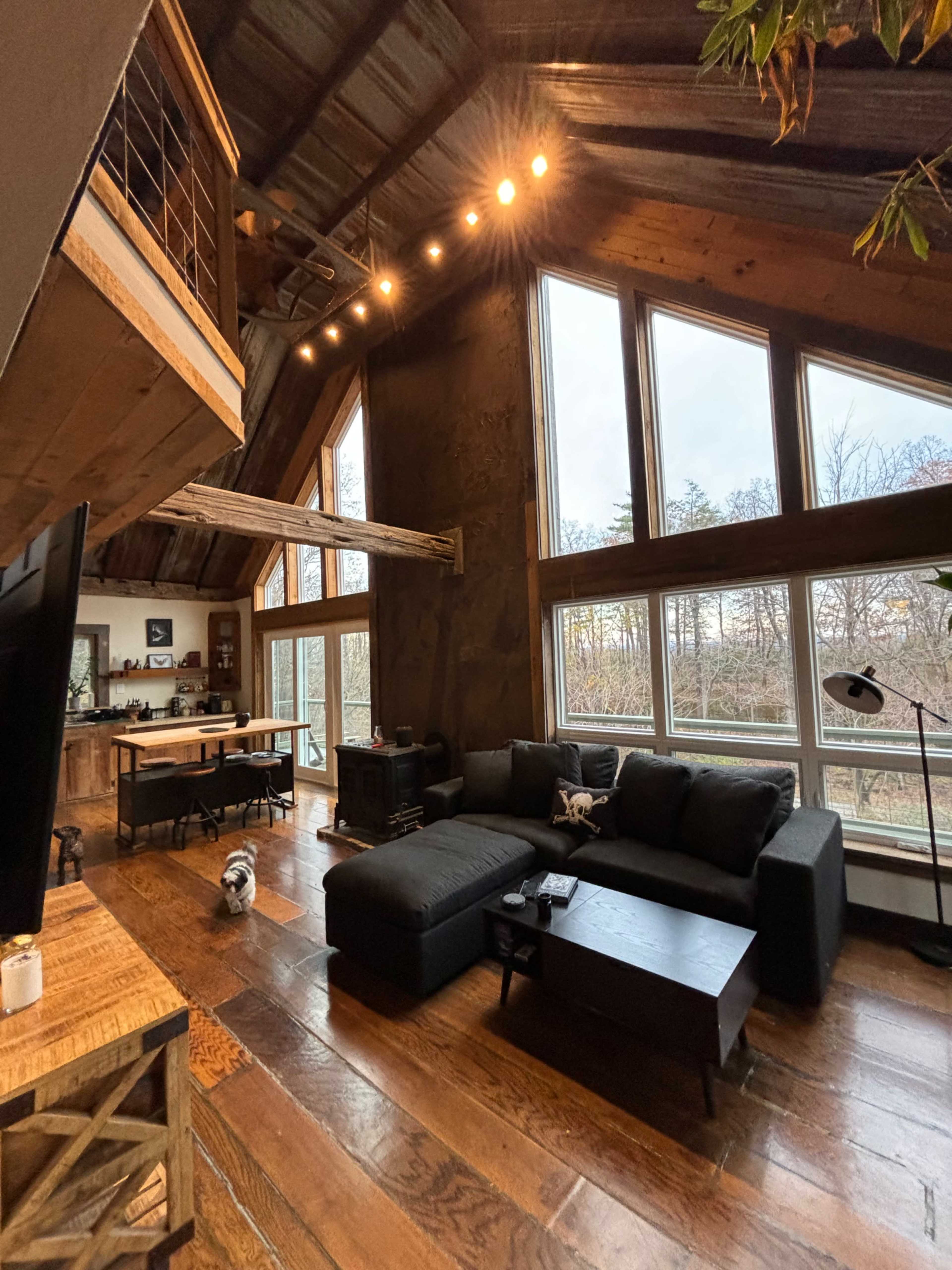 The image shows a cozy interior of a cabin featuring a dark sectional sofa, wooden floors, and large windows that offer a view of the outdoors.
