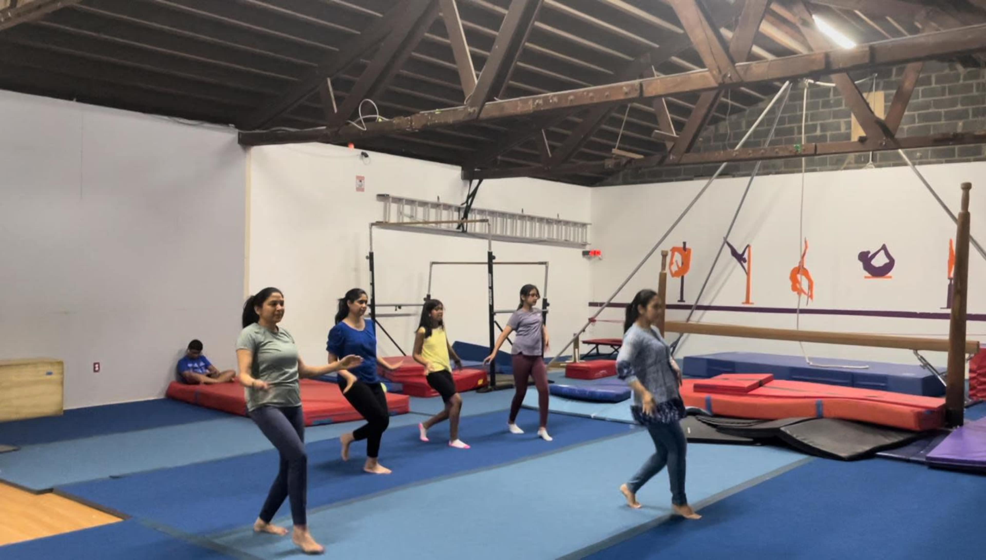 A group of six women practices gymnastics on mats in a gymnasium with various equipment in the background.