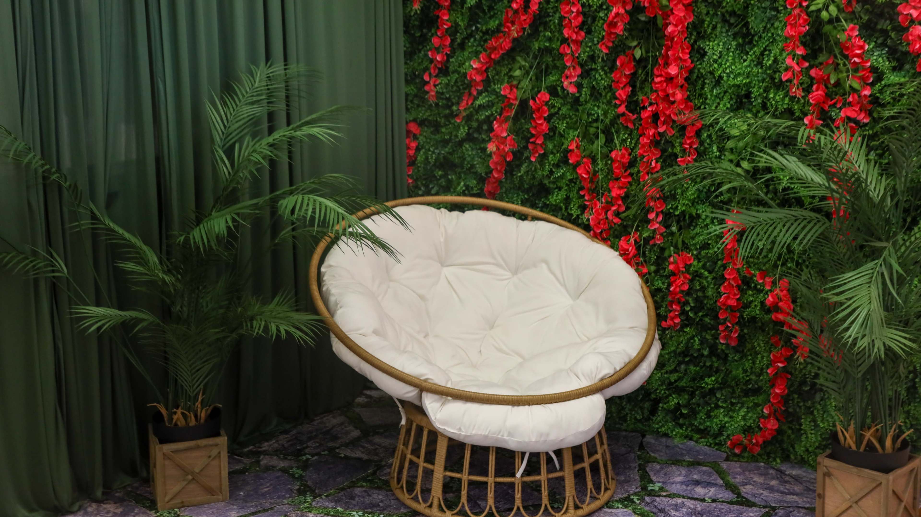 A white, cushioned chair with a rattan base is positioned in front of a green backdrop adorned with red flowers and decorative plants.