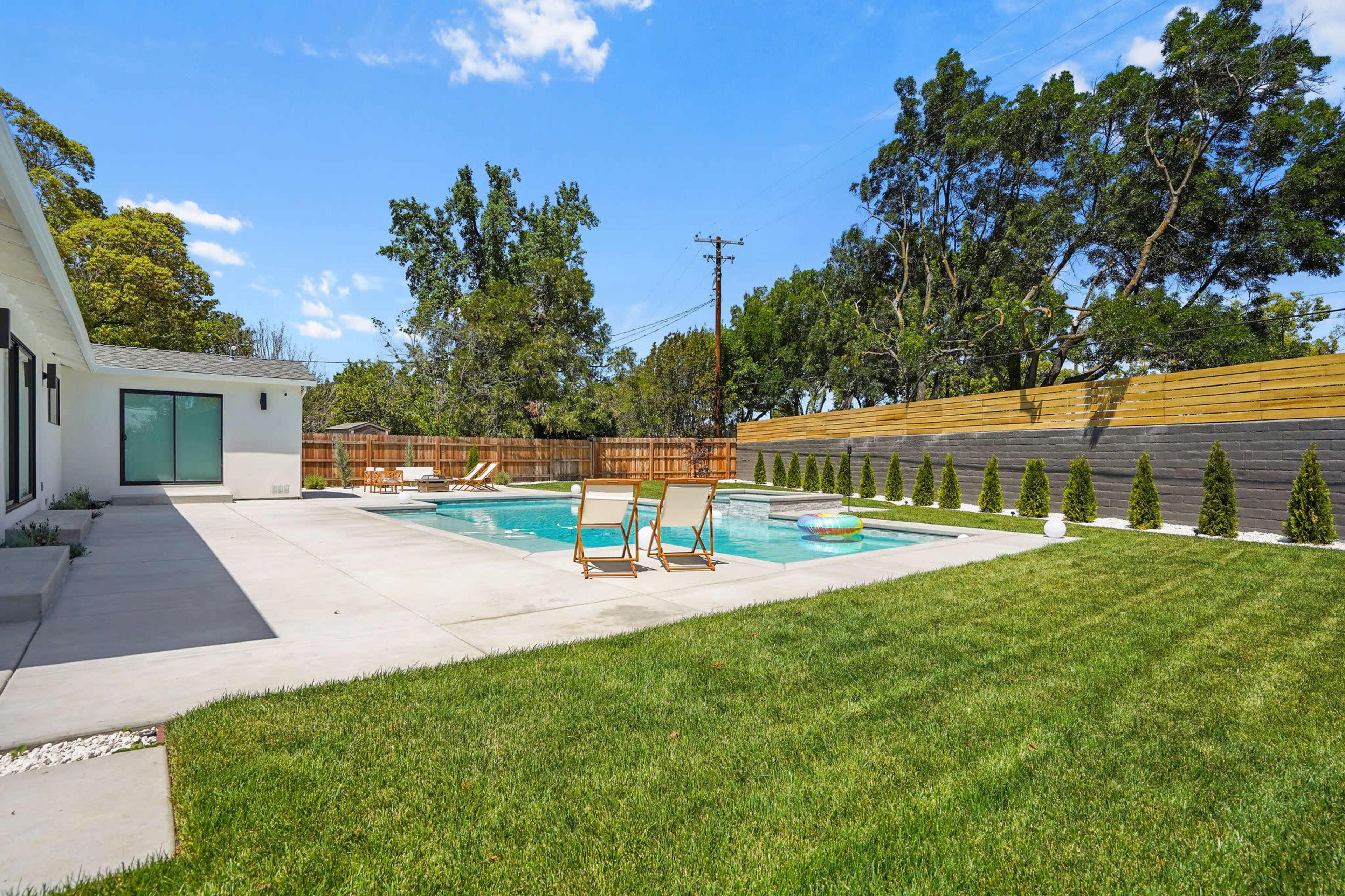 A patio area features a swimming pool surrounded by green grass and lounge chairs, with trees and wooden fencing in the background.
