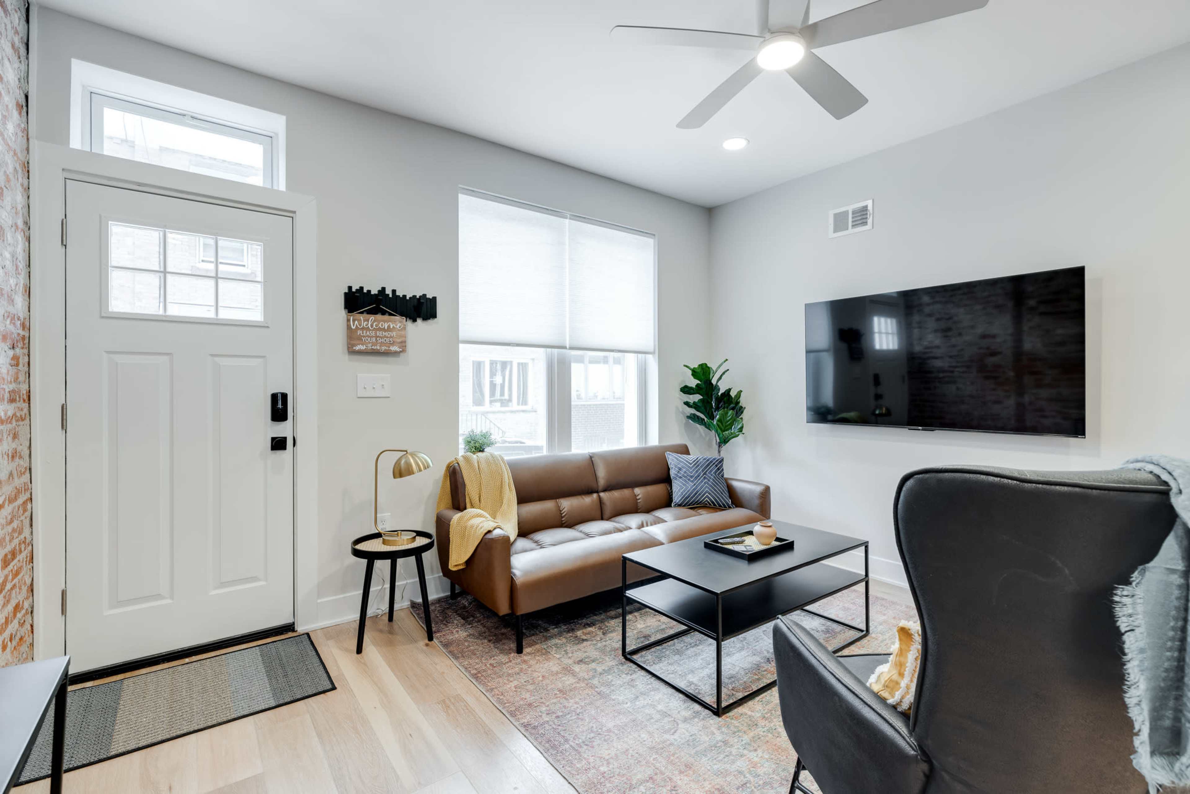A modern living room with a brown sofa, a coffee table, and a television mounted on the wall, featuring a large window and a front door.